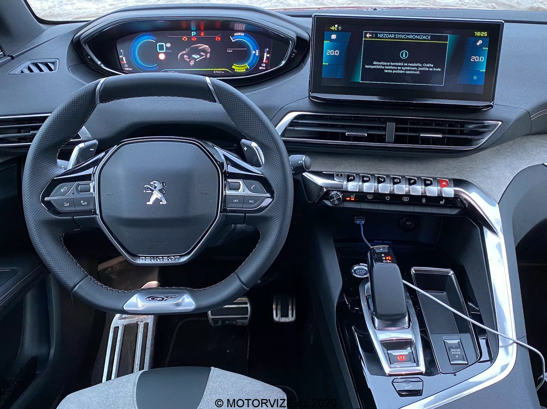 Vista interior de un Peugeot 3008 de 2021 desde la perspectiva de los asientos traseros que muestra la cabina delantera: volante multifuncional moderno con diseño de fondo plano y botones de control, panel de instrumentos digital, consola central elevada con selector de marchas y freno de mano electrónico, sistema de infoentretenimiento con pantalla táctil independiente, rejillas de ventilación rectangulares con controles de clima, diseño de interiores elegante con material similar a cuero, acentos metálicos, acabado negro brillante, conectividad USB, indicaciones de transmisión automática, portavasos y espacio de almacenamiento.