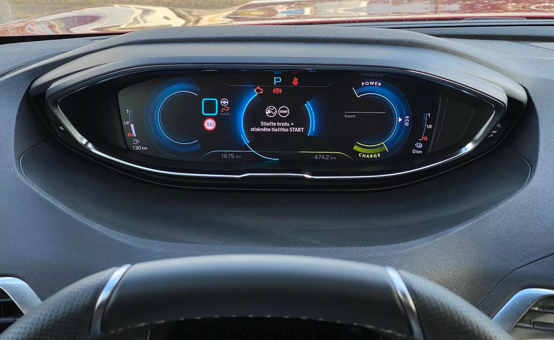 Vista del conductor del panel de instrumentos del Peugeot 3008 de 2021 con un salpicadero totalmente digital que muestra las métricas e indicadores del vehículo. A la izquierda, un indicador de potencia sugiere que el vehículo es eléctrico/híbrido; en el centro superior, advertencias de cinturón y airbag; a la derecha, el velocímetro que indica que el vehículo está estacionario; en la parte inferior se muestra el cambio de marcha (parking), el odómetro, la temperatura exterior, modos de conducción y autonomía disponible. Volante de tres radios parcialmente visible. Vista desde detrás del volante con un ligero ángulo hacia abajo.