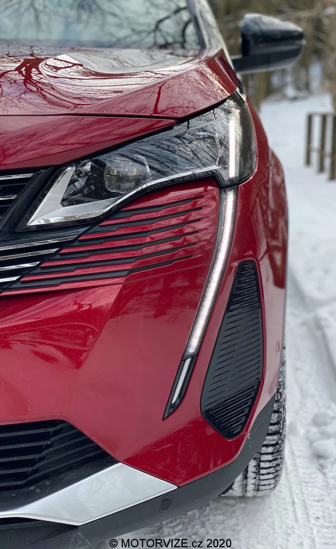 Vista de cerca en ángulo bajo de un moderno Peugeot 3008 2021 rojo. Se enfoca en el cuarto frontal izquierdo que muestra faros LED angulares elegantes, una parrilla grande y agresiva, un diseño de ventilación pronunciado, acabados de pintura brillantes y paneles de carrocería contorneados. El espejo lateral tiene señales de giro integradas, y la escena incluye suelo nevado, sugiriendo condiciones invernales. Marca de agua de MOTORVIZE.cz visible.