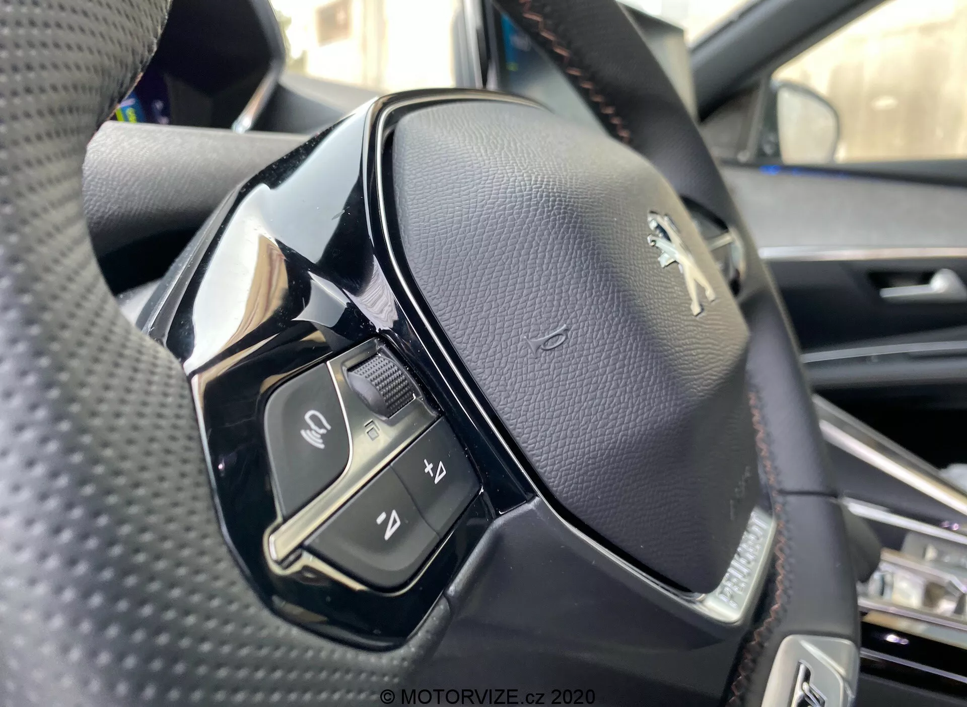 Vista de primer plano de un volante desde la perspectiva del asiento del conductor en el Peugeot 3008 2021. La imagen enfatiza el lado izquierdo del volante, tomada desde un ángulo ligeramente bajo y capturando los detalles de los controles del lado izquierdo y el logo de Peugeot. El volante está recubierto de cuero negro perforado con una mezcla de materiales negros mate y brillantes. Los controles visibles incluyen un interruptor, botones para funciones de medios y comunicación como aceptar o finalizar llamadas, activación de control por voz y otra función no identificada. El enfoque es nítido, mostrando las costuras en el cuero y el acabado brillante en algunas partes de plástico, posiblemente botones táctiles.