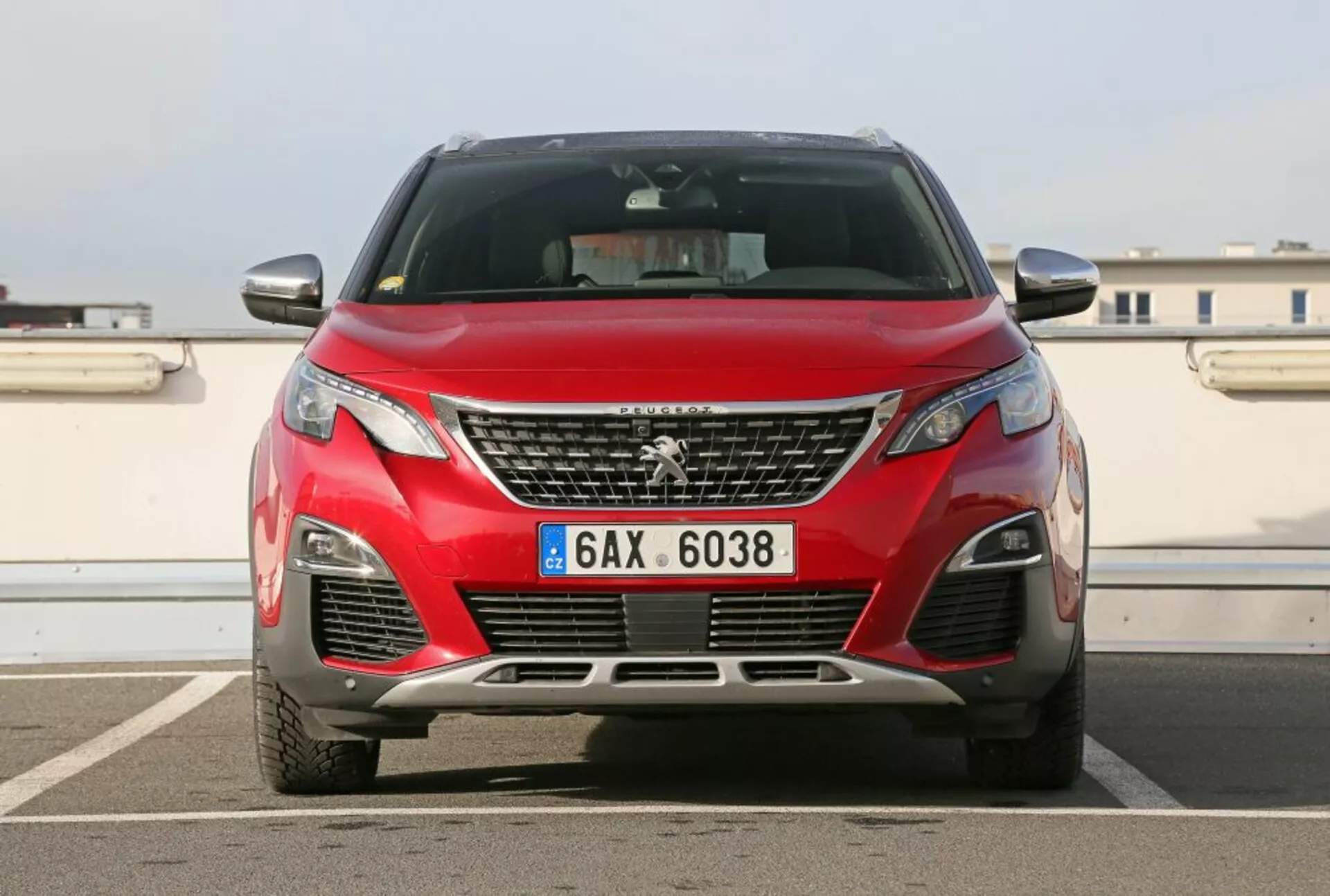Vista frontal de un Peugeot 3008 rojo del 2016 mostrando el emblema en la parrilla, faros angulares, rejillas de ventilación inferior con luces antiniebla, sugerencia de los contornos del capó contra un fondo urbano con cielo despejado