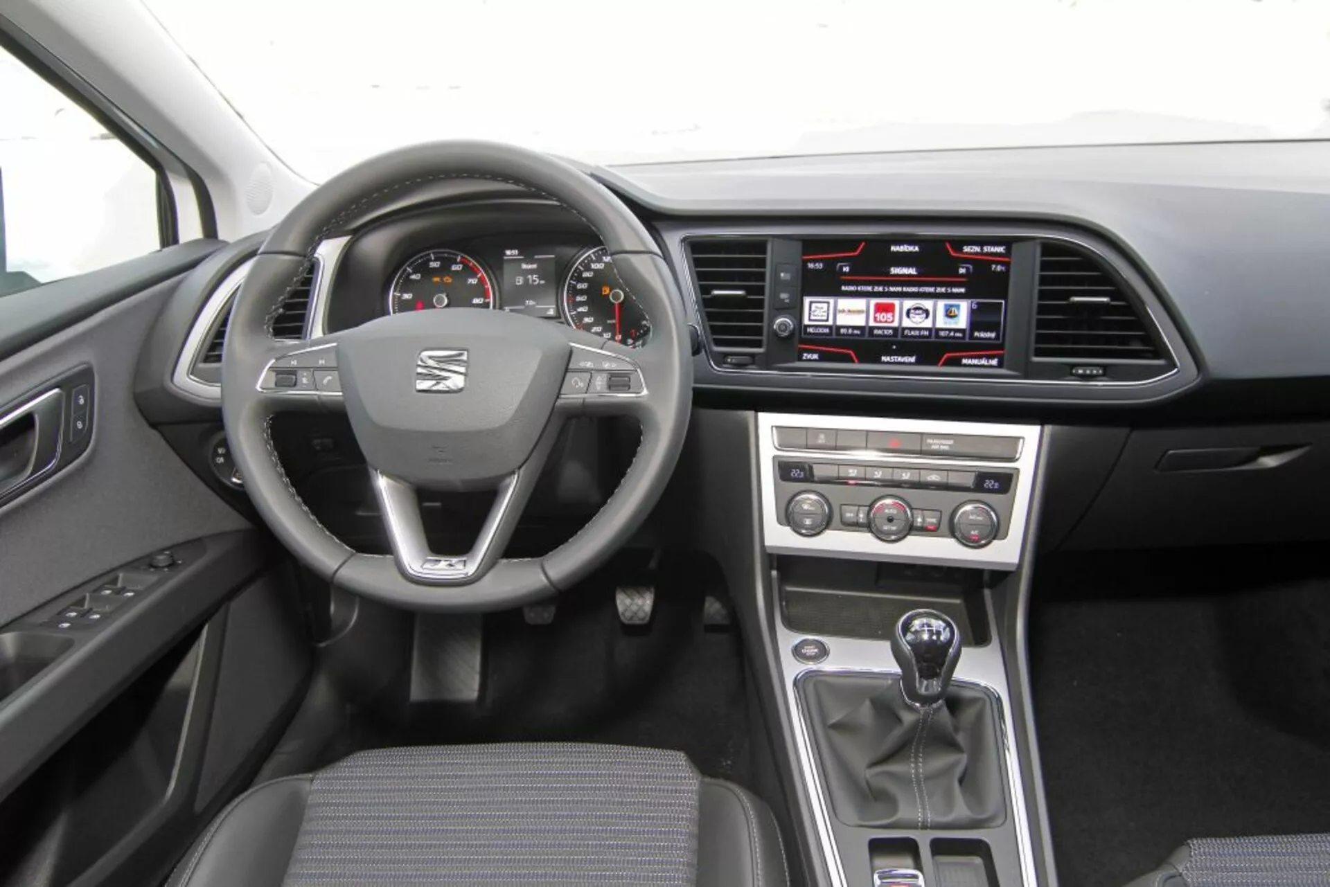 Vista interior de un Peugeot 308 SW de 2013 mostrando el volante con logo y controles multifunción, velocímetro y tacómetro analógicos con pantalla digital, sistema de infoentretenimiento táctil en el tablero, botones de control manual del clima en la consola central, palanca de cambios manual y tapicería práctica de tela oscura.