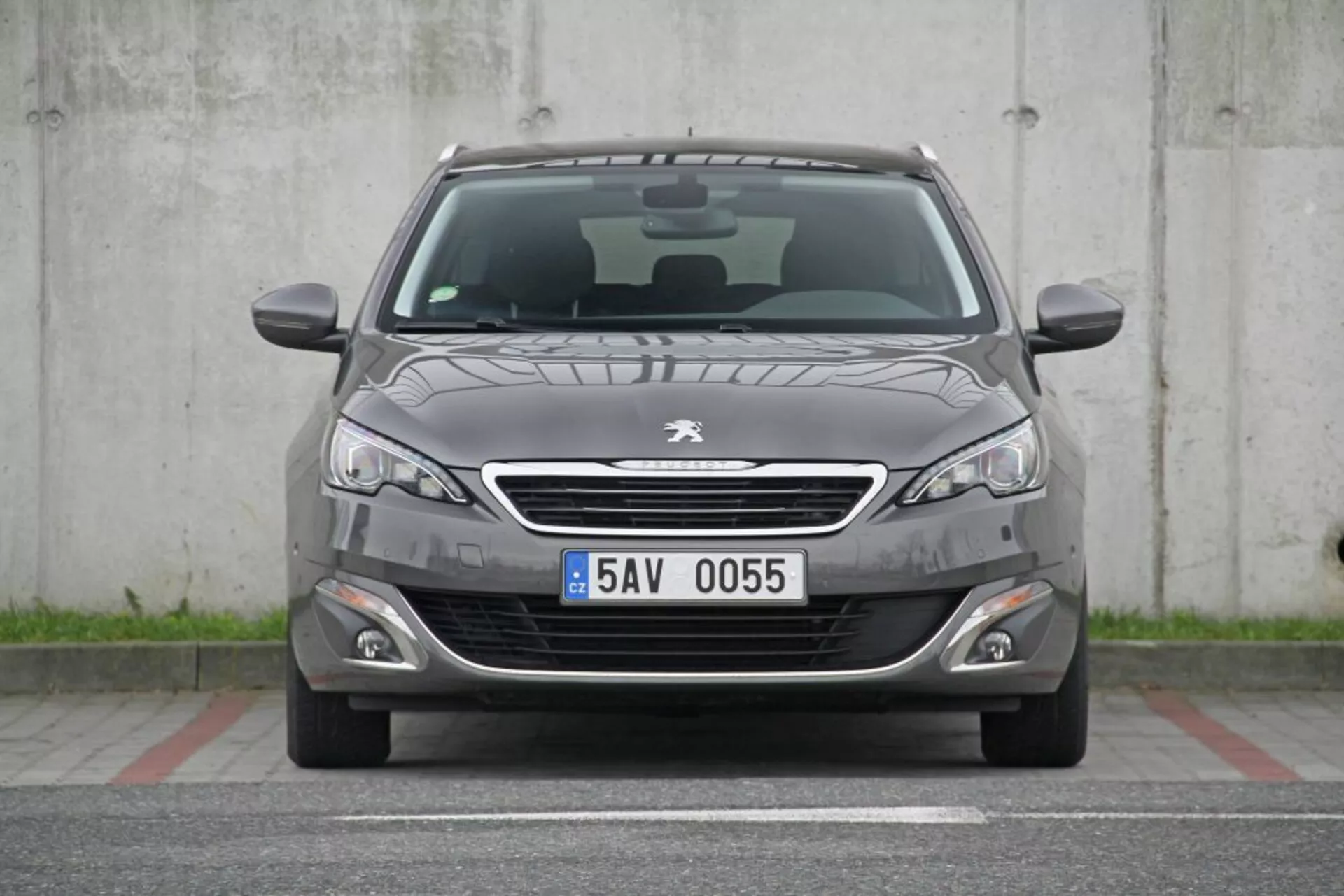 Vista frontal de un Peugeot 308 SW de 2013 en gris oscuro con parrilla cromada, faros elegantes, luces antiniebla, paragolpes delantero deportivo, suaves pliegues en el capó, limpiaparabrisas visibles, placa de matrícula europea, sensores de aparcamiento, rejilla inferior de panal y techo panorámico visible, estacionado en un área de hormigón sin obstrucciones