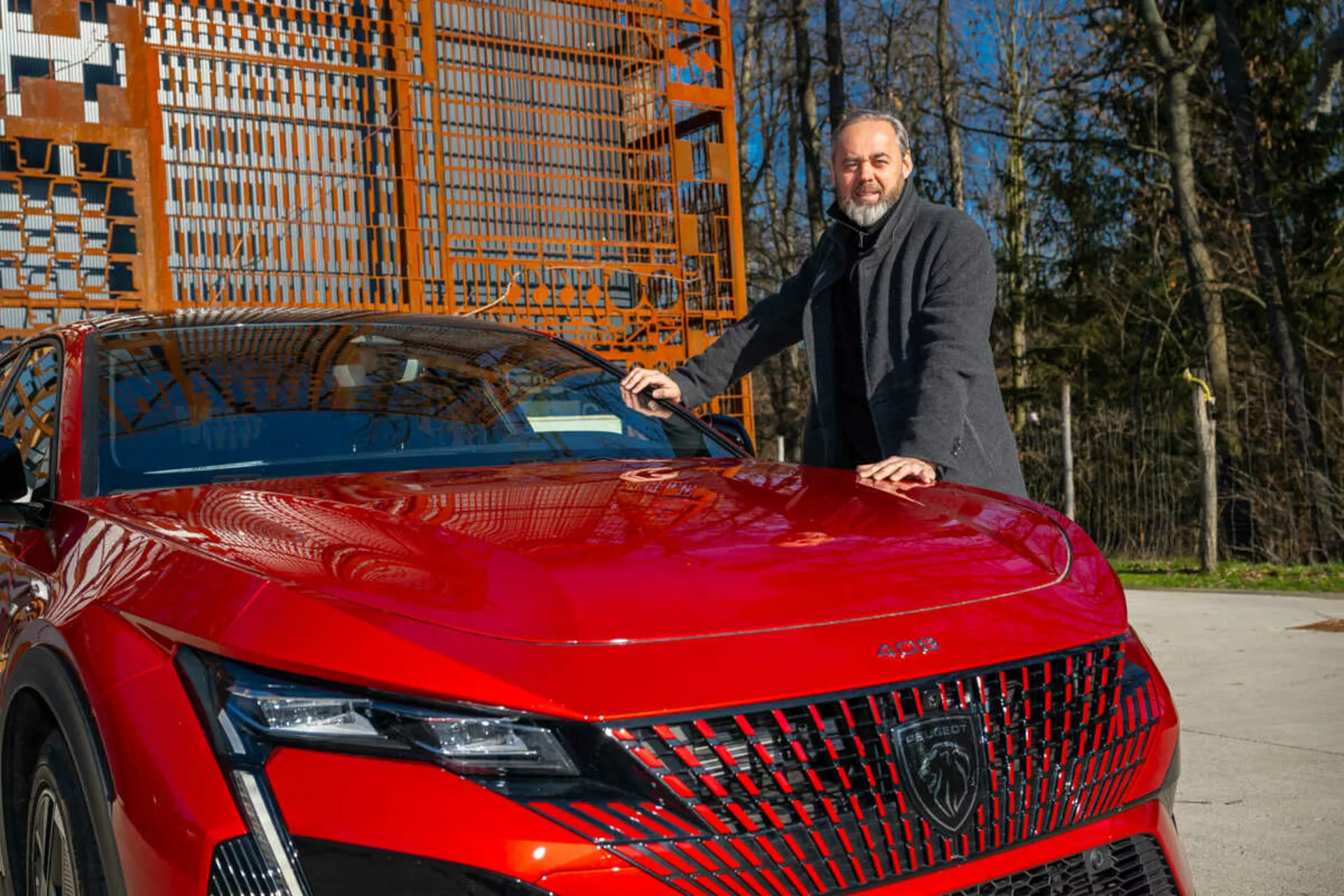 Modelo rojo del Peugeot 408 de 2023 mostrando su cuarto delantero izquierdo a la luz del día, destacando los faros LED detallados, la parrilla geométrica y el capó deportivo y aerodinámico con un hombre en un abrigo oscuro a su lado colocando su mano en el capó