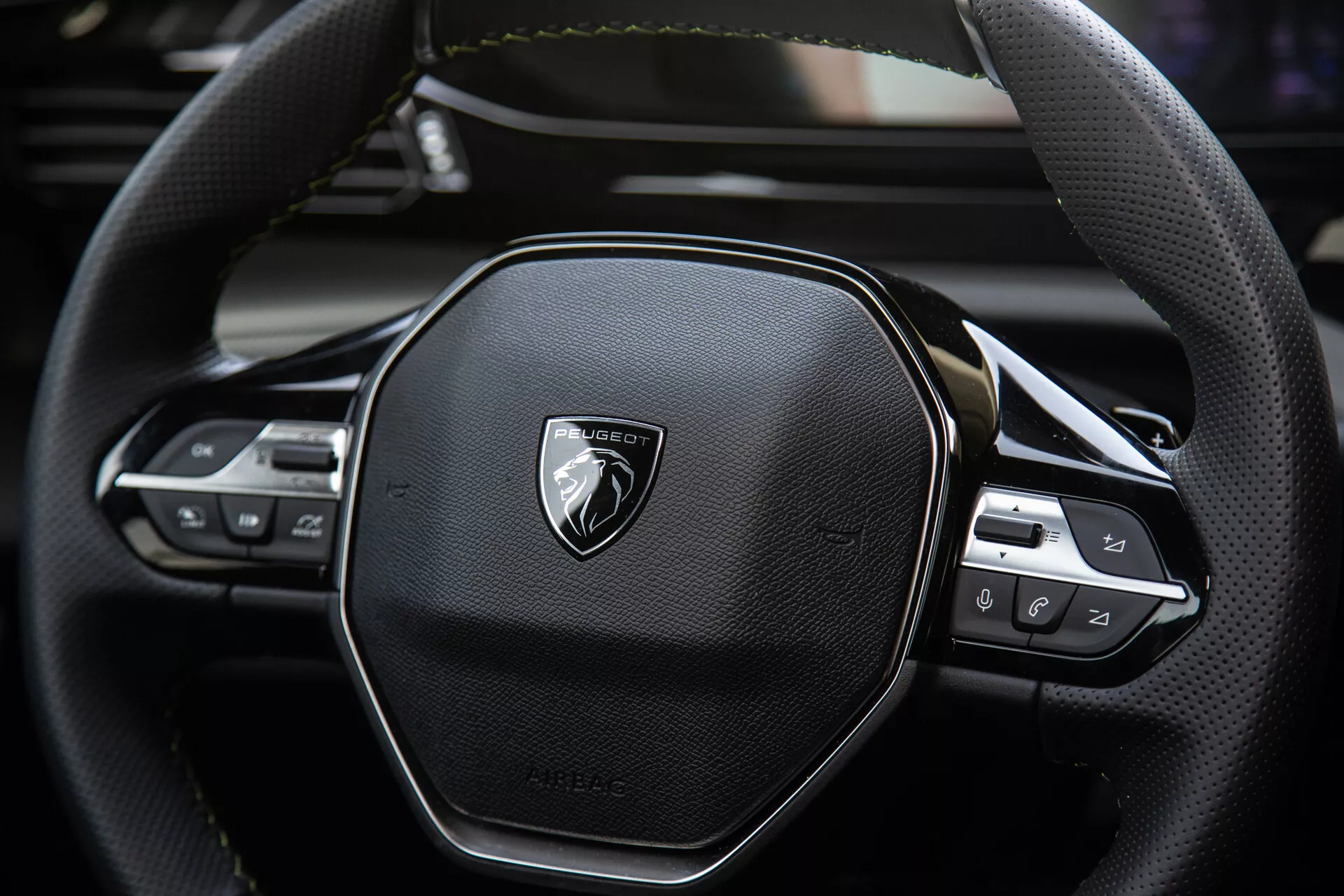 Vista cercana del volante del Peugeot 408 2023 que muestra diseño ergonómico hexagonal, costuras de cuero, controles tecnológicos integrados para audio y configuraciones de crucero, con emblema en metal cepillado.