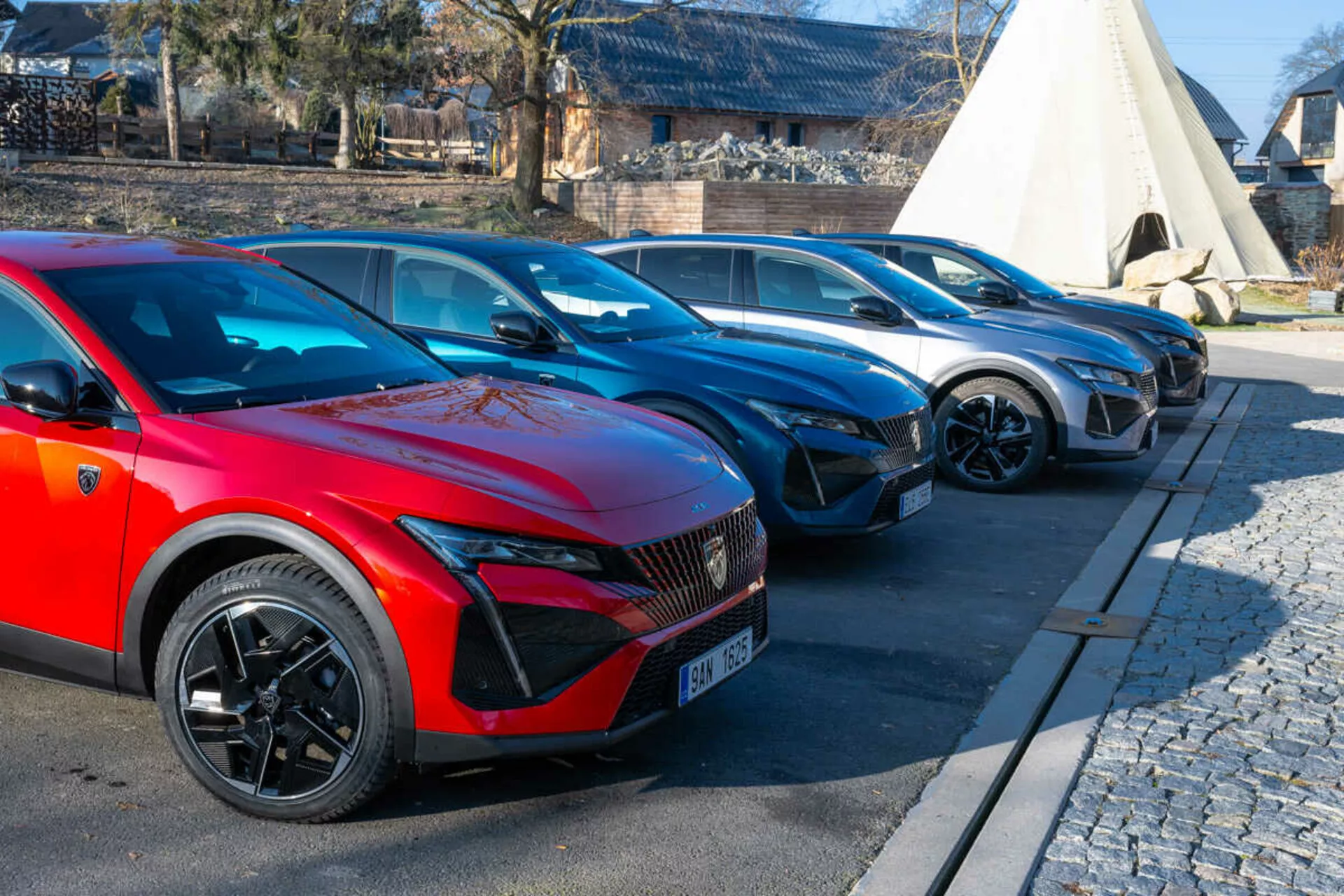 Alineación de Peugeot 408 de 2023 con coche rojo en primer plano con ruedas de múltiples radios deportivos y faros LED indicativos de modelos eléctricos o híbridos, aparcado en área pavimentada junto a empedrado