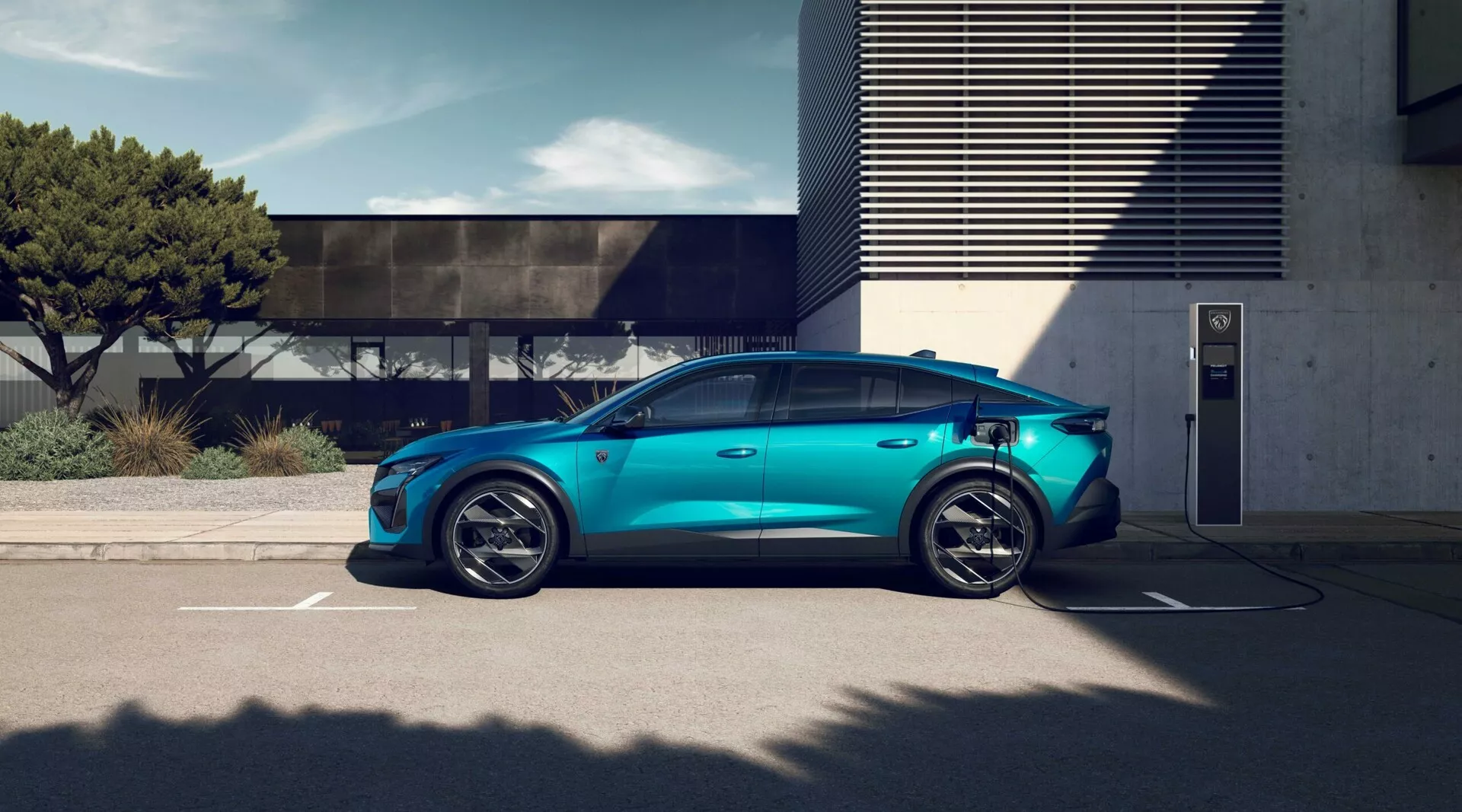 Vehículo eléctrico Peugeot 408 de 2023 en azul metálico aparcado en estación de carga exterior, mostrando llantas de aleación de cinco radios deportivas, silueta aerodinámica, manijas de puertas empotradas, puerto de carga visible, espejos laterales convencionales, ventanas con tintado de privacidad y techo solar panorámico bajo condiciones de luz diurna brillante con arquitectura moderna al fondo