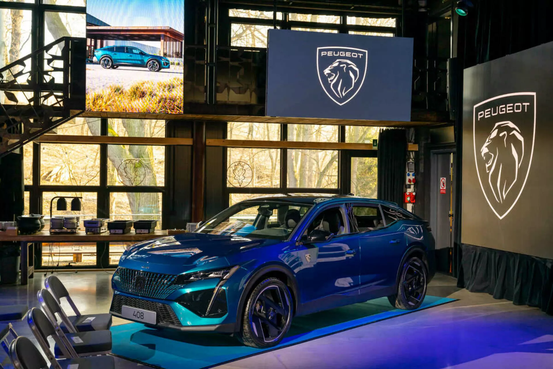 Peugeot 408 azul del 2023 exhibido en un evento interior, destacando una parrilla distintiva, faros LED agudos, capó deportivo contorneado con entradas de aire, ruedas de aleación grandes y un perfil lateral dinámico con intermitentes integrados en los espejos laterales montados en la puerta