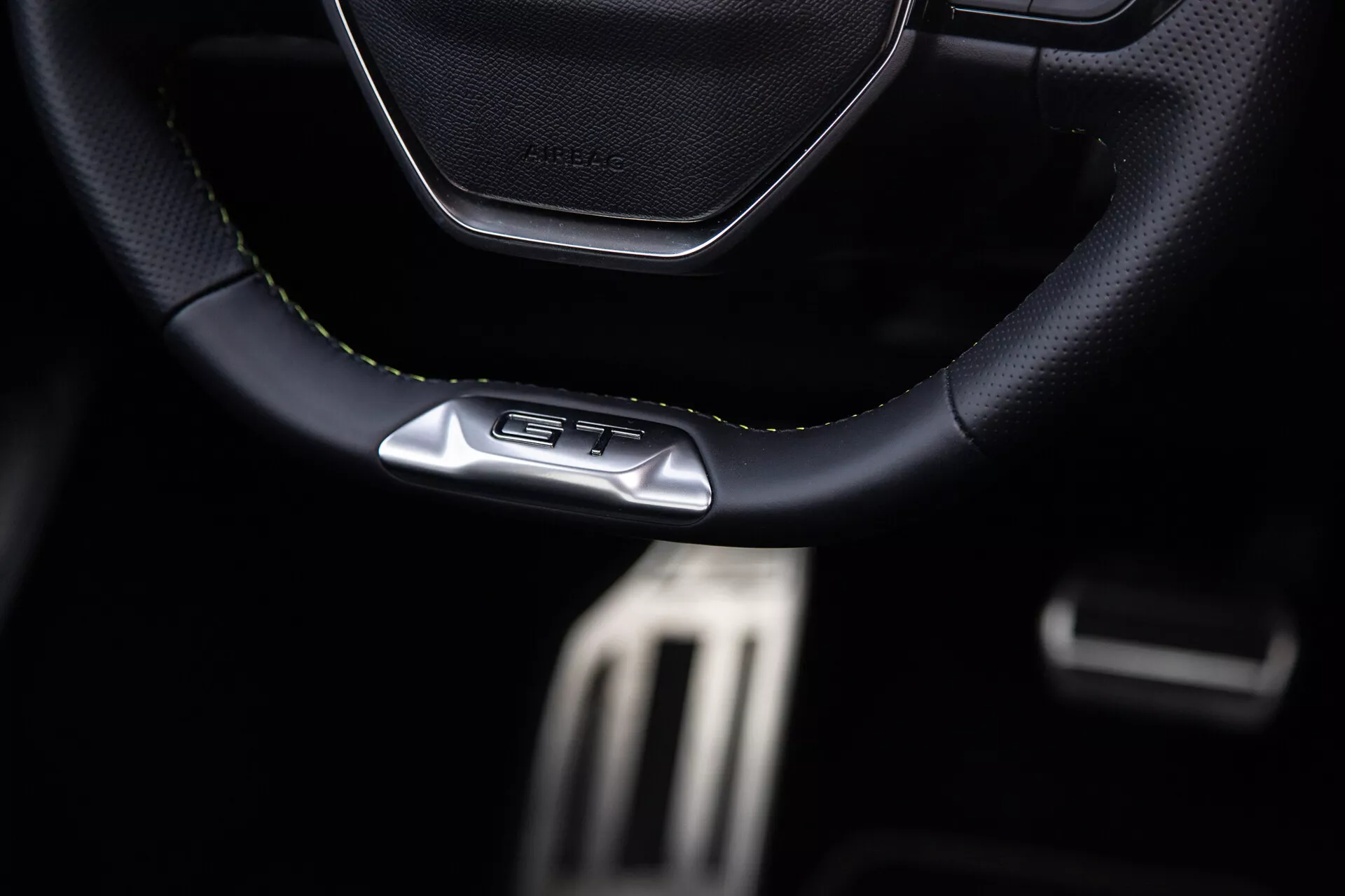 Interior del Peugeot 408 2023 centrado en el volante deportivo con costuras de cuero con emblema GT, pedales metálicos de rendimiento y características de cabina oscura premium