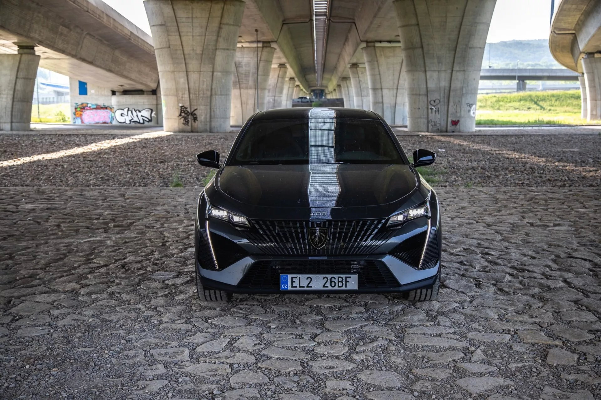 Peugeot 408 2023 en una vista frontal dinámica que muestra su postura agresiva y diseño deportivo con parrilla detallada, faros LED elegantes y características aerodinámicas en un pavimento estampado bajo un puente urbano