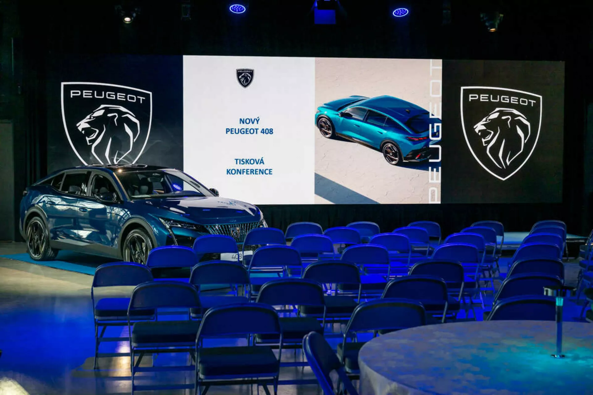 Peugeot 408 exhibido en una conferencia de prensa, visto en un ángulo de 45 grados destacando sus perfiles frontal y lateral, diseño elegante y faros LED, con pantallas digitales y branding en el fondo