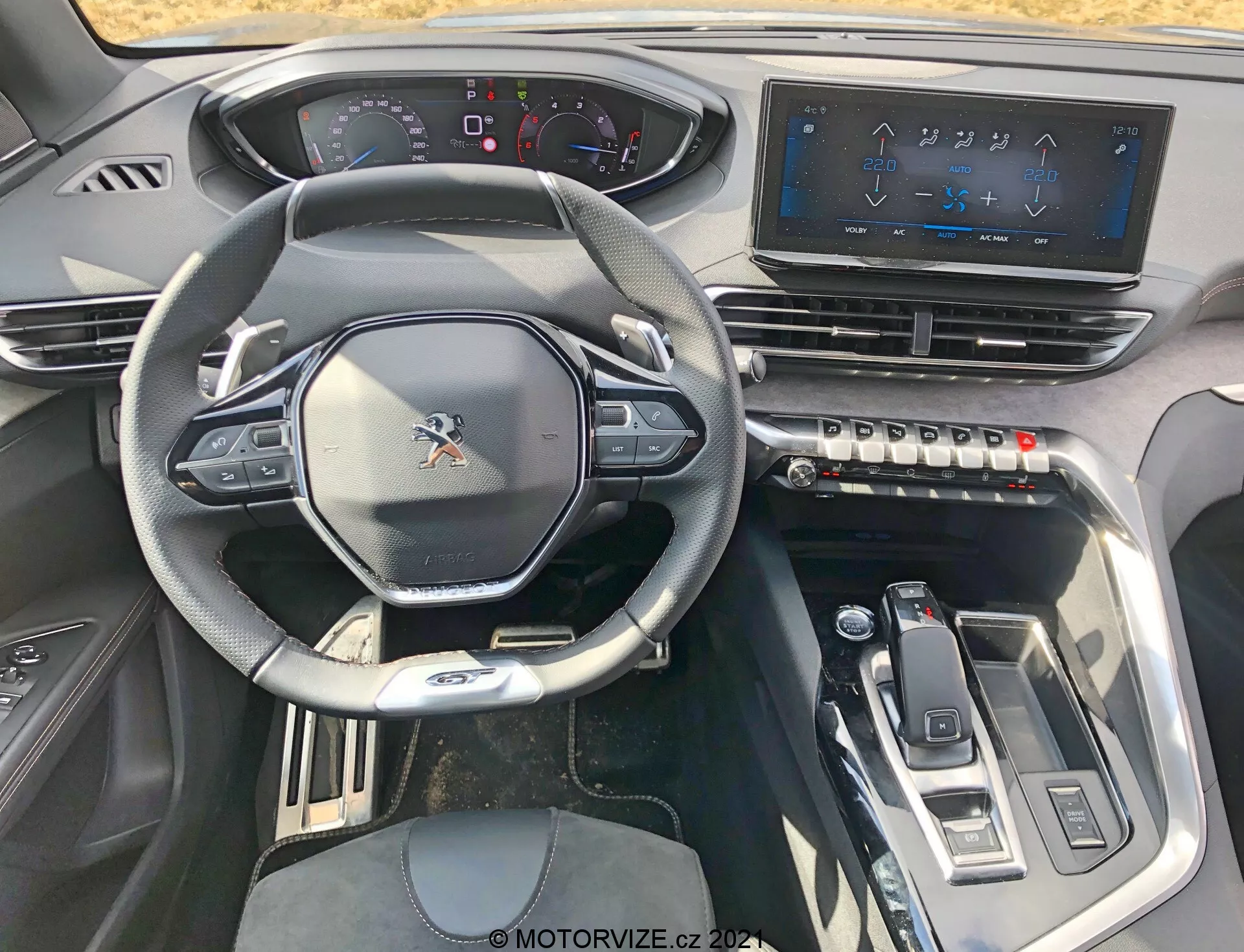 Vista interior de un Peugeot 5008 2021 desde el asiento del conductor, que muestra el volante multifunción deportivo con controles integrados, un panel de instrumentos parcialmente digital, un gran sistema de infotainment con pantalla táctil, botones táctiles para el control del clima, un selector de marchas elegante y elementos de diseño moderno con acentos metálicos para una experiencia tecnológica y de alta gama.