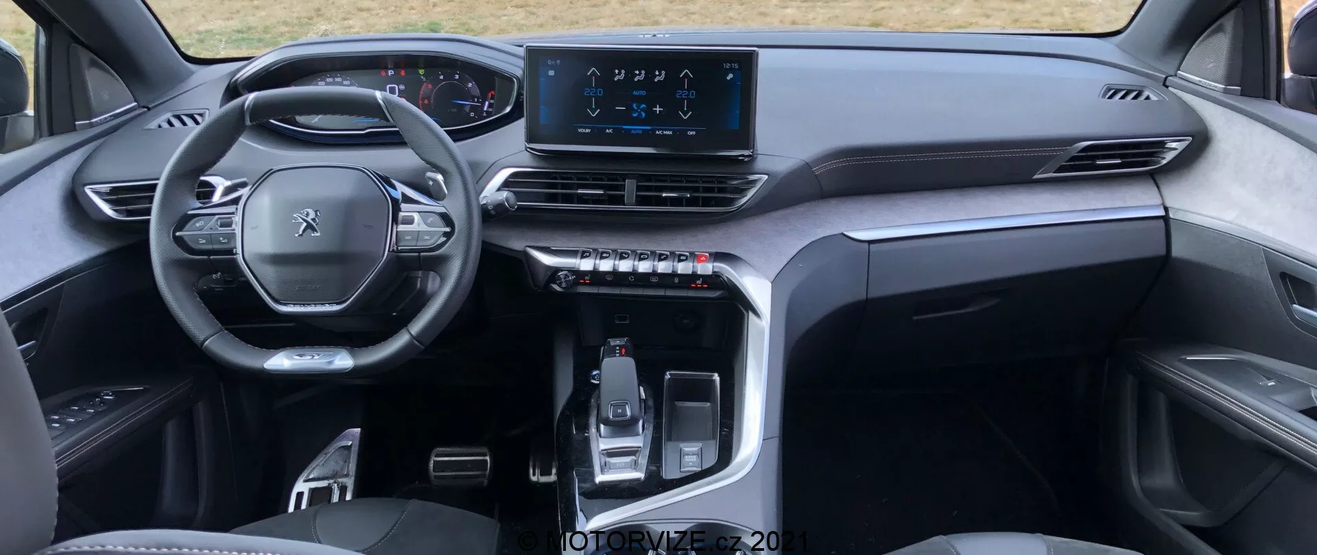 Vista interior del Peugeot 5008 2021 desde la perspectiva del asiento trasero, destacando un volante multifuncional, un panel de instrumentos digital, un tablero simétrico con ventilaciones de aire, una pantalla táctil de infoentretenimiento central y una elegante consola central con una palanca de cambios controlada electrónicamente y botones táctiles. El diseño minimalista y elegante utiliza materiales de alta calidad, sugiriendo un vehículo moderno y premium.