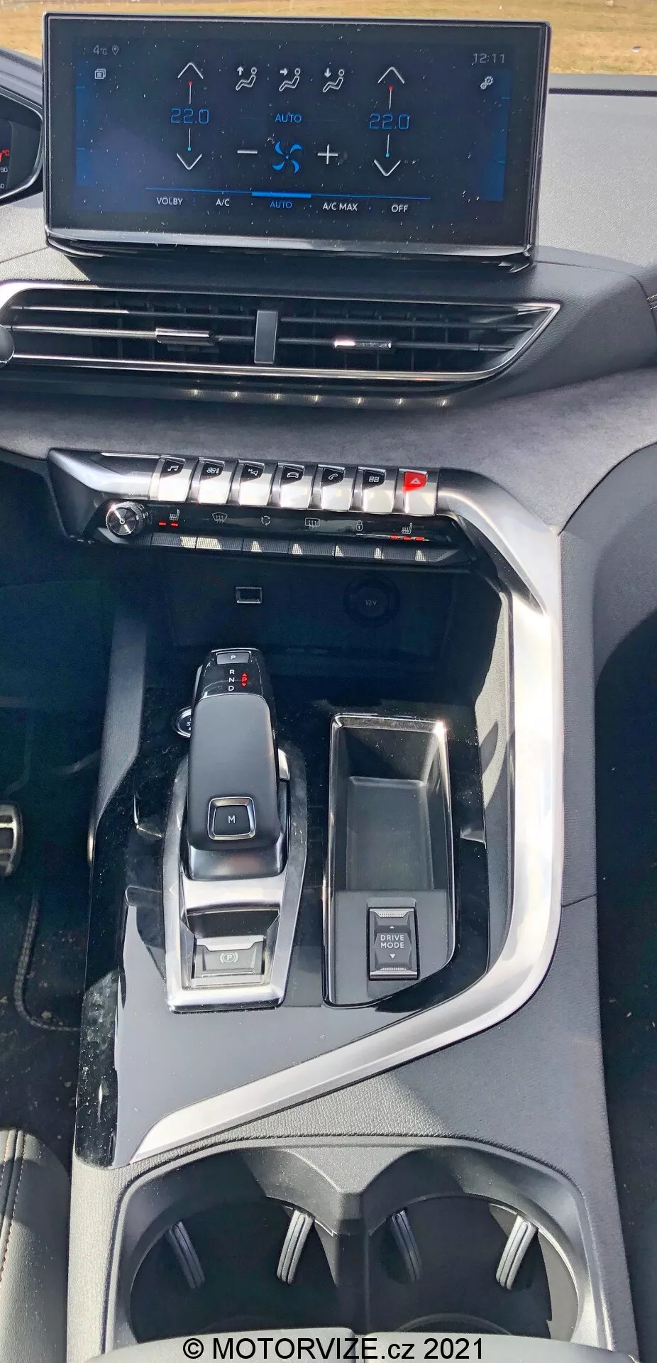 Vista interior del Peugeot 5008 de 2021 que muestra la consola central desde un ángulo superior. La imagen destaca una pantalla táctil flotante de infoentretenimiento que muestra los controles de aire acondicionado sobre botones y diales físicos para el control del clima. Más abajo se observa una palanca de cambios electrónica y elegante, junto con controles circundantes para la selección de modos de conducción y un mando de interfaz multifuncional. El diseño indica un vehículo moderno y premium con características tecnológicas avanzadas y ergonómicas, incluidos los portavasos y espacios de almacenamiento.