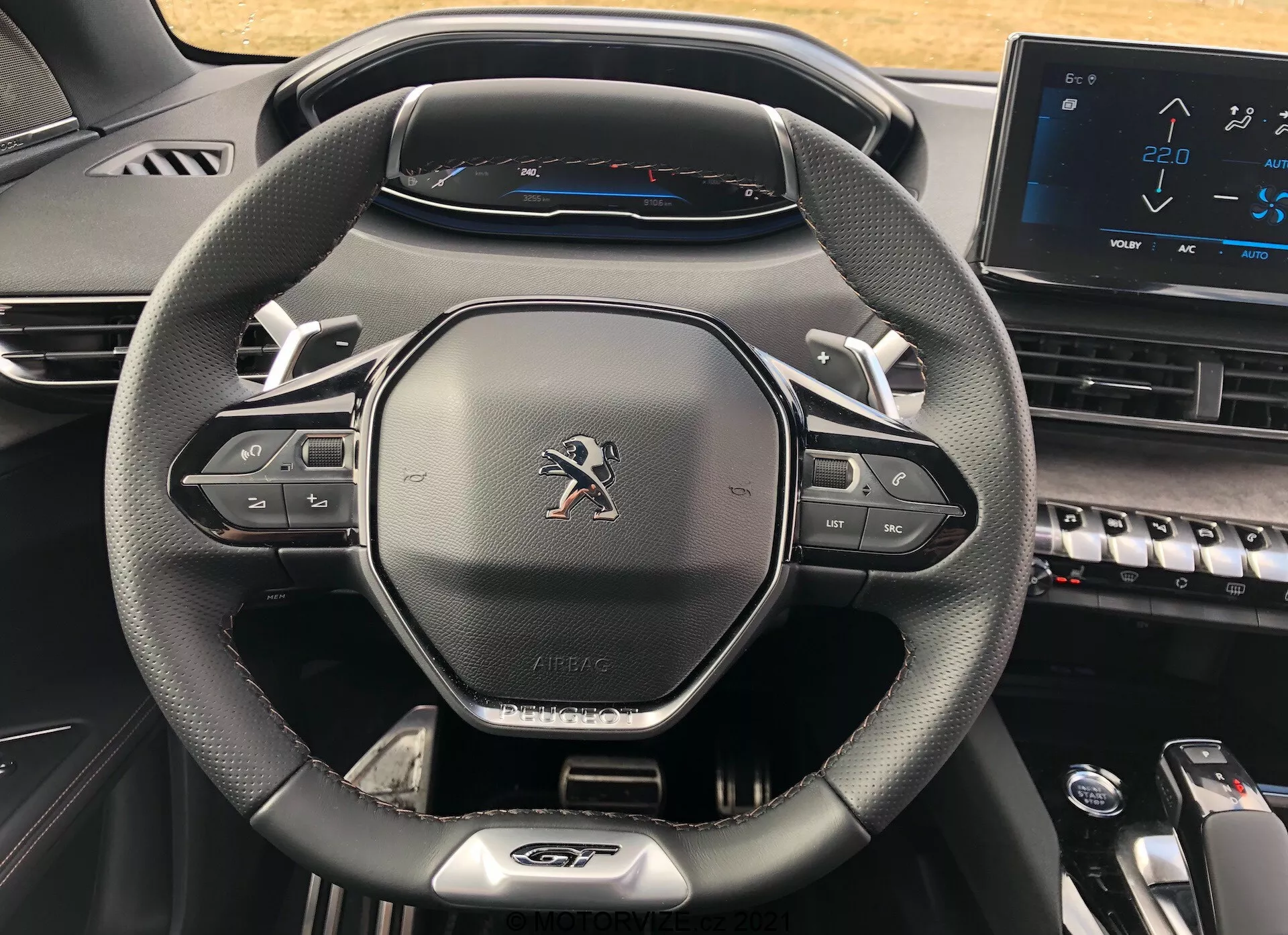 Vista en primer plano del habitáculo del conductor dentro de un Peugeot 5008 2021, destacando el volante moderno de tres radios con textura de cuero, botones de control multimedia y de crucero, emblema de Peugeot, rótulo 'AIRBAG', cuadro de instrumentos digital completamente iluminado que muestra un velocímetro con una velocidad máxima de 220 km/h y parte de la consola central con teclas de estilo piano y una pantalla táctil electrónica, que muestra un diseño contemporáneo y tecnológicamente avanzado.