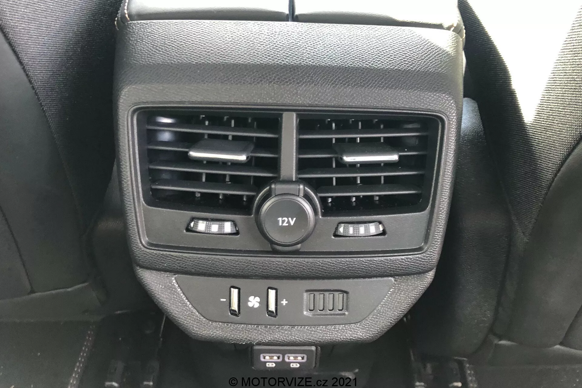 Vista de cerca de las características para pasajeros traseros en la parte trasera de la consola central en el Peugeot 5008 2021. La imagen muestra dos salidas de aire, una toma de corriente de 12V, controles de calefacción de asientos, puertos USB y un botón en blanco, capturada desde la perspectiva de un ocupante del asiento trasero.