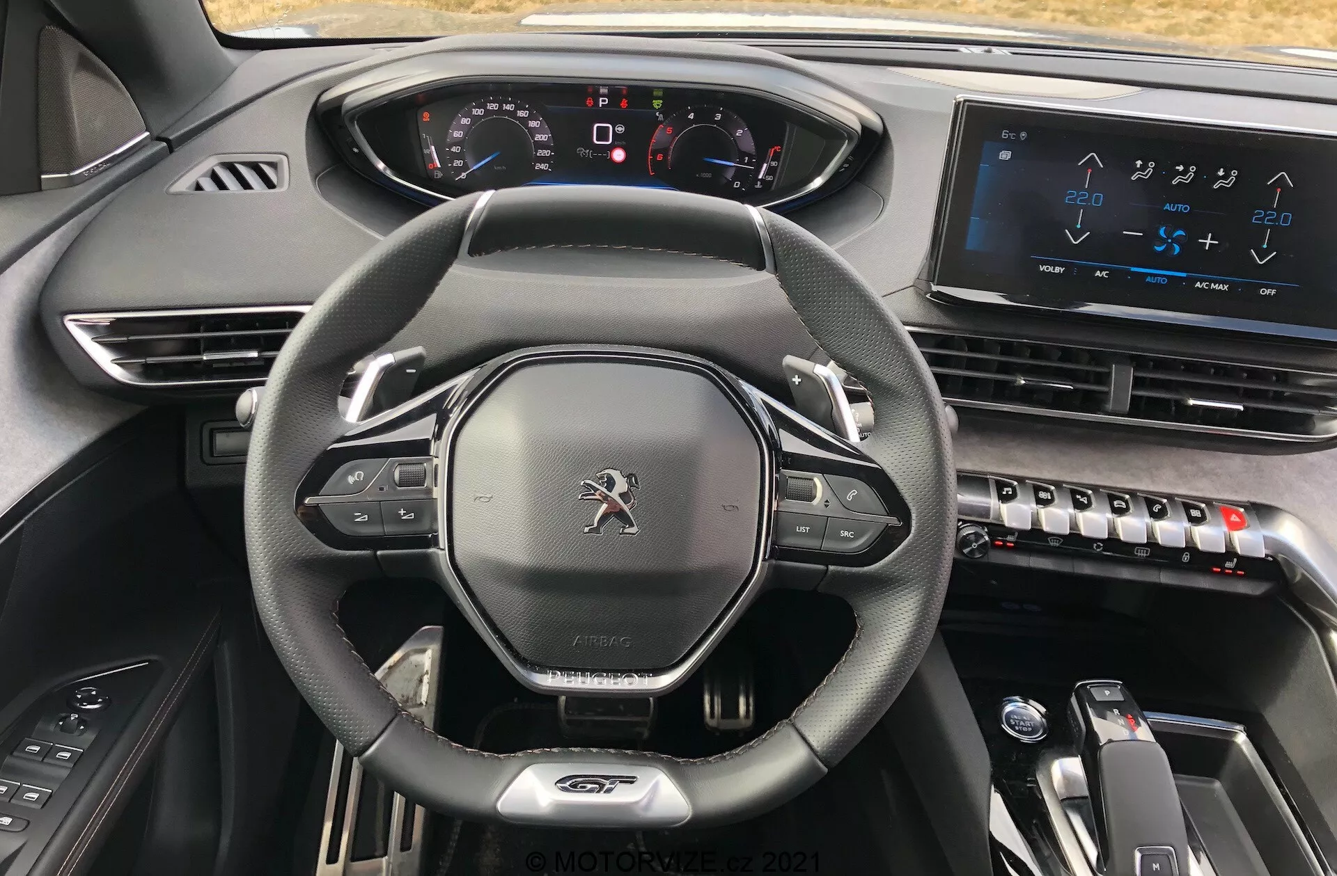 Vista interior del Peugeot 5008 2021 desde el asiento del conductor, destacando el volante con revestimiento de cuero de fondo plano con controles integrados, un panel de instrumentos totalmente digital con indicadores iluminados, una gran pantalla táctil de sistema de infoentretenimiento en la consola central, controles de aire acondicionado elegantes, selector de marchas electrónico para transmisión automática y un diseño de materiales oscuros de alta calidad que transmite una estética moderna, avanzada y premium.