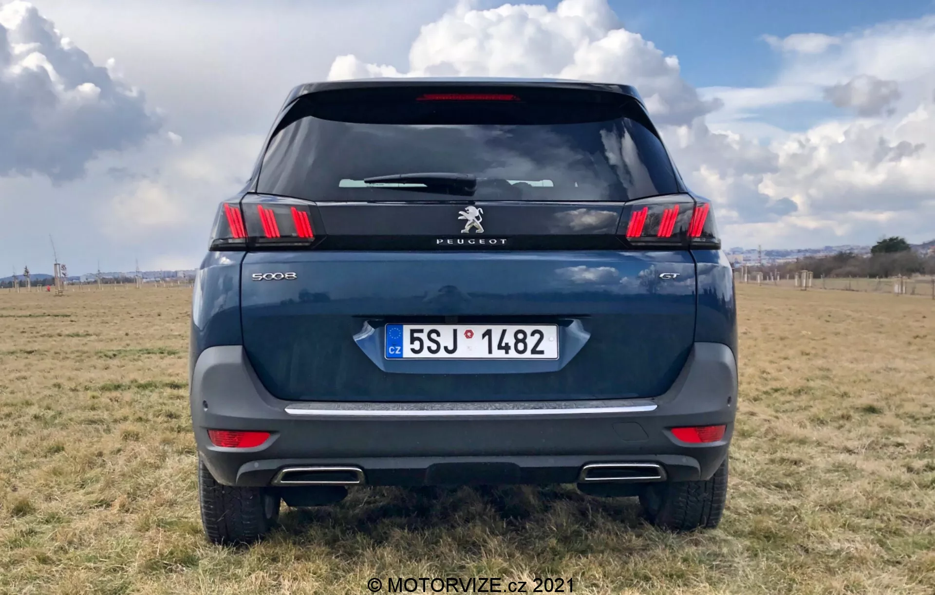 Vista trasera de un moderno SUV Peugeot 5008, resaltando las elegantes luces traseras horizontales con segmentos LED en forma de garra, el logo de Peugeot y la inscripción del modelo '5008'. Color azul oscuro o gris, estacionado en un terreno al aire libre con un cielo parcialmente nublado. Los elementos visibles incluyen parabrisas trasero, limpiaparabrisas trasero, lámpara de freno de montaje alto, manija del portón trasero y placa de matrícula de la República Checa con 'CZ'. Se aprecian la tira reflectante sobre el parachoques con sensores de aparcamiento y los recortes de punta de escape. Foto centrada con el coche llenando la mayor parte del marco.