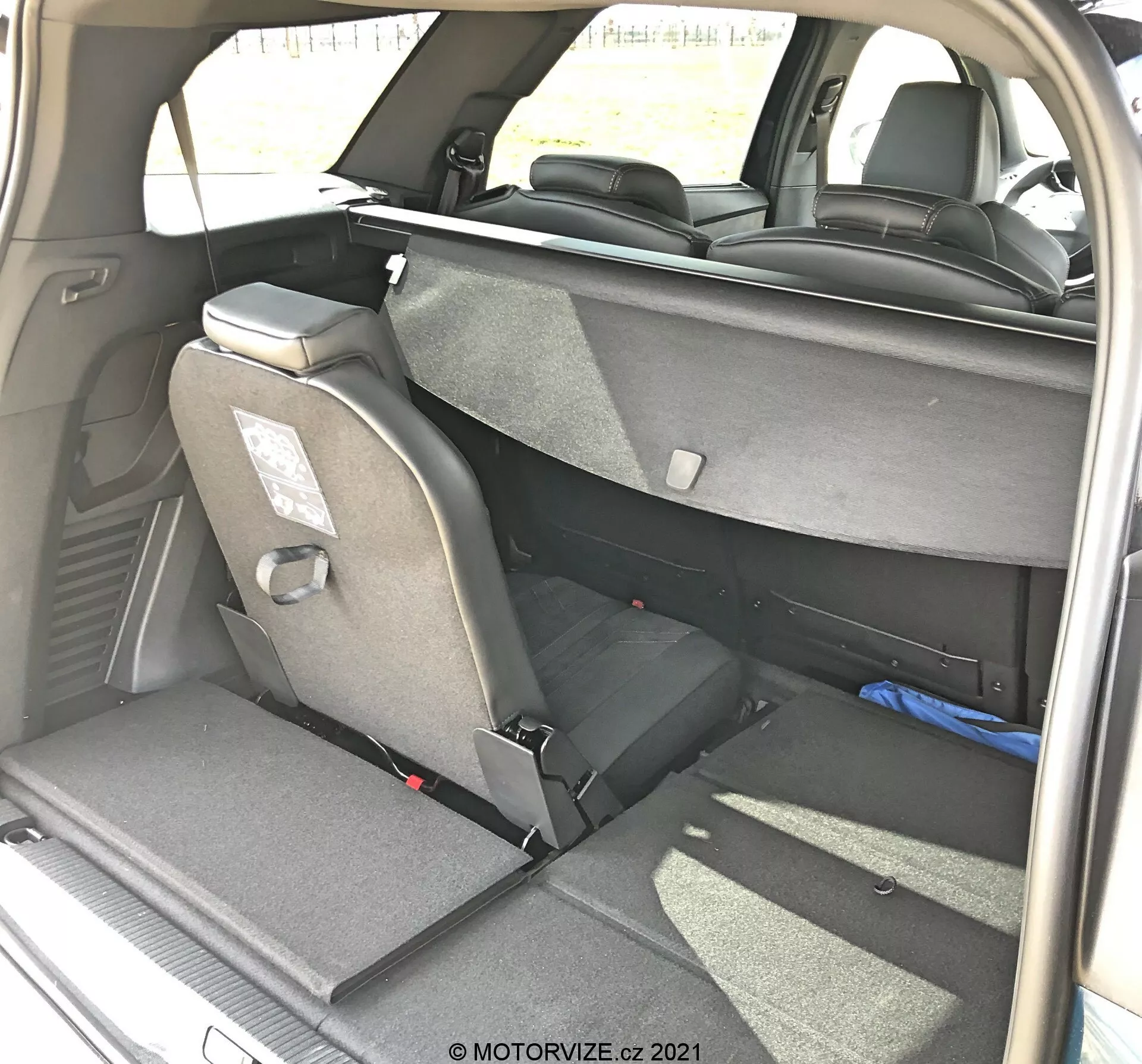 Vista interior del área de carga de un Peugeot 5008 2021 desde un ángulo trasero derecho de tres cuartos, mostrando una disposición de asientos flexible con los asientos traseros parcialmente plegados, tapicería de tela, reposacabezas y cinturones de seguridad. Asiento trasero derecho completamente plegado, asiento izquierdo en posición vertical, indicando funcionalidad de plegado dividido 60/40. Interior de color oscuro visible junto con iluminación diurna natural, tomada por MOTORVIZE.cz.