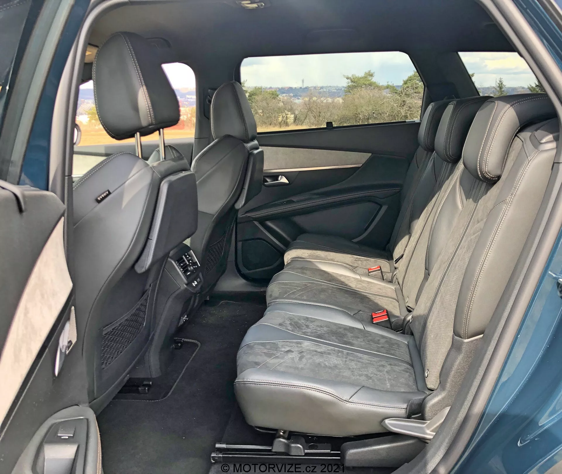 Interior de un Peugeot 5008 2021 desde la perspectiva trasera derecha mostrando el compartimento de pasajeros trasero. La puerta trasera derecha está abierta revelando características del panel de la puerta como manija, almacenamiento y altavoz. Se pueden ver tres asientos traseros tapizados en cuero gris con reposacabezas. El reposabrazos del asiento central está en posición vertical. También se ven cinturones de seguridad traseros y asientos delanteros con reposacabezas ajustables y consola central. Respiraderos de control de clima traseros en la consola central, ventana derecha tintada, vislumbres de la puerta trasera izquierda que indican simetría. En general, interior bien mantenido sin funciones tecnológicas visibles.