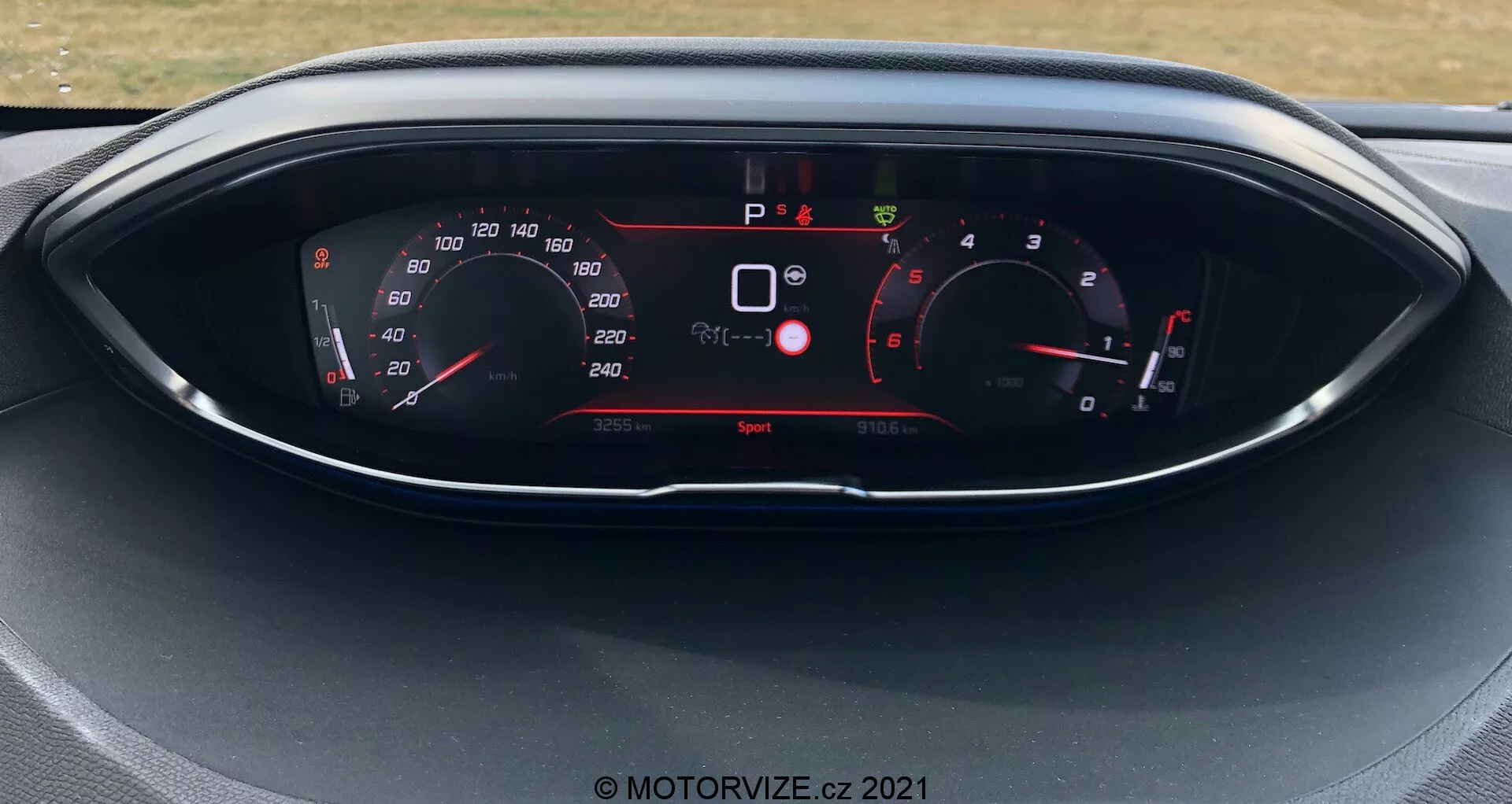 Vista de cerca del cuadro de instrumentos en un Peugeot 5008 de 2021, con una pantalla digital y medidores de estilo analógico. La pantalla muestra velocímetro, tacómetro, selección de marchas e indicadores varios del vehículo como advertencias de seguridad y modo de conducción. El coche está estacionado con el modo de conducción 'Sport' activado.