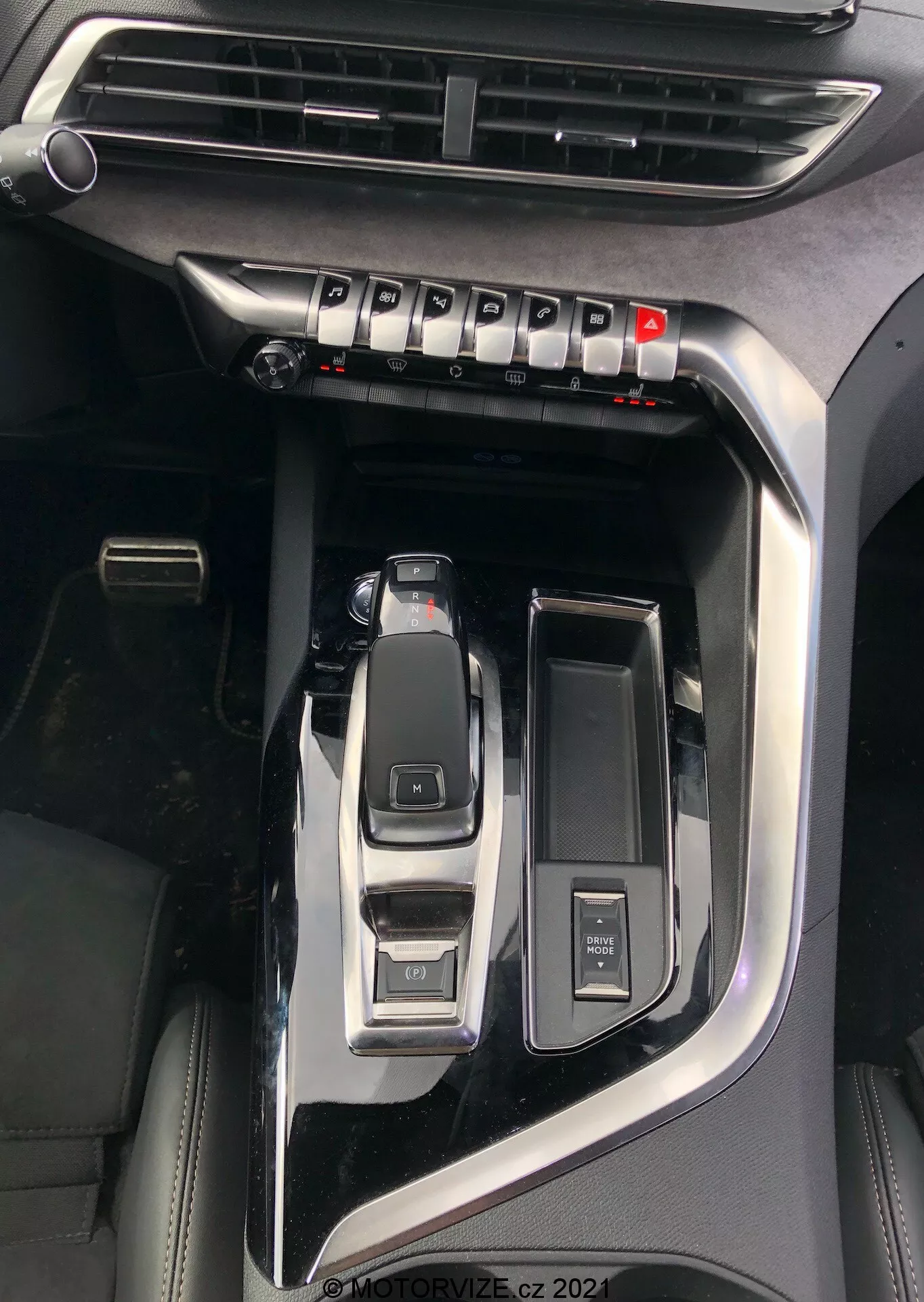 Vista de cerca de la consola central del Peugeot 5008 2021 destacando características clave como la palanca de cambios automática en posición de aparcamiento, el selector de modos de conducción para modos eco, sport, comfort, un botón de encendido/parada para el arranque sin llave, un mando giratorio de control multimedia, un portavasos abierto, una rejilla de ventilación visible para el control del clima y un acabado en negro piano con acentos metálicos que indican un modelo premium, marca de agua 'MOTORVIZE.cz 2022'.