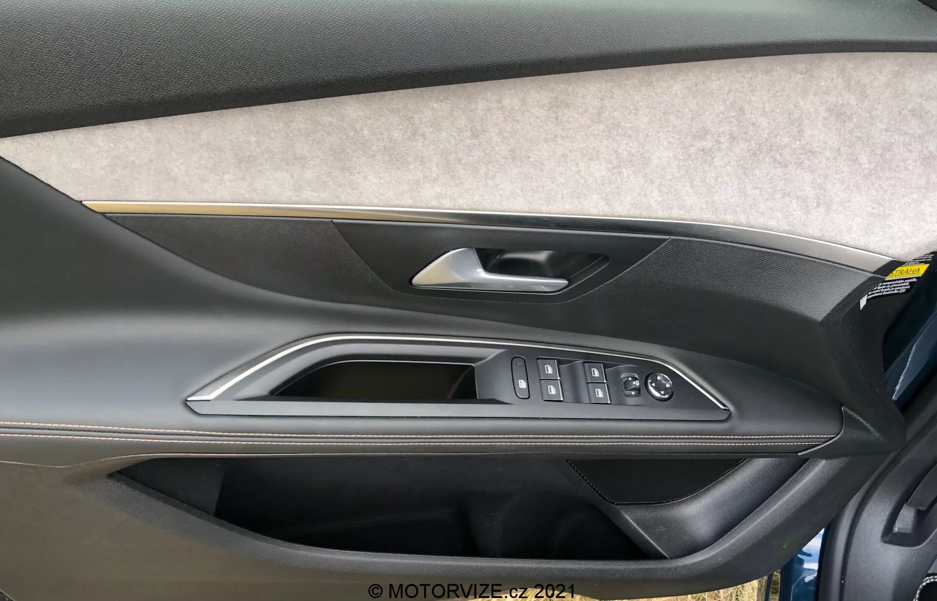 Vista de primer plano del panel de la puerta interior del Peugeot 5008 2021 que muestra una sección tapizada de fieltro o tela, un diseño de plástico moldeado de dos tonos con una porción superior más oscura y una inferior más clara, una manija de puerta metálica o cromada de alta calidad, un adorno pulido y controles ergonómicos para las ventanas eléctricas, cierre de puertas y espejos laterales. También se ve un bolsillo de almacenamiento en la parte inferior del panel de la puerta.
