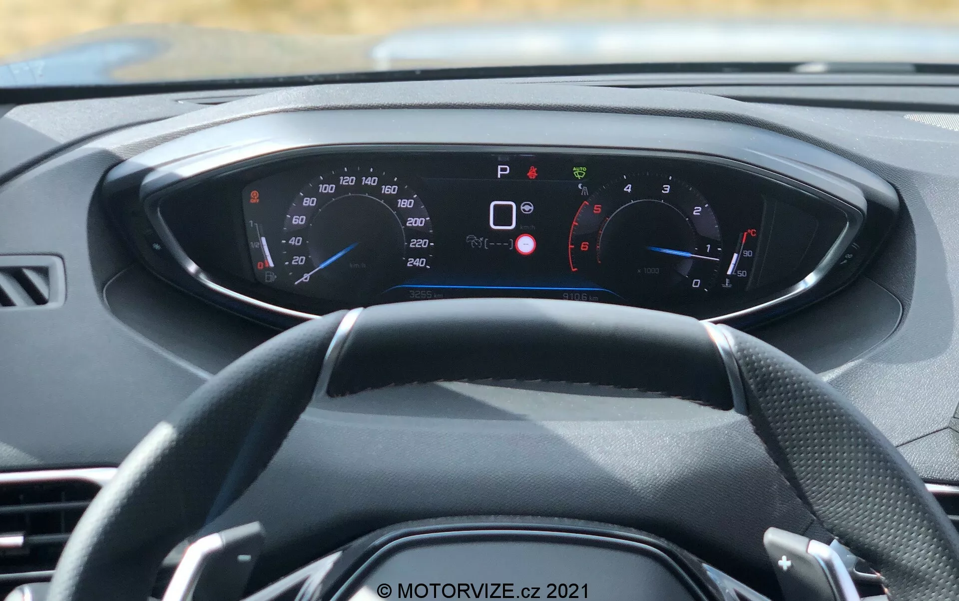 Vista interior de un vehículo moderno desde una perspectiva de primera persona, enfocándose en el volante y el tablero digital. El volante de tres radios está centrado, con varios medidores e indicadores en el tablero iluminado detrás de él. El velocímetro y el tacómetro son visibles, con lecturas en km/h y mph, adecuado para mercados internacionales. El tablero muestra numerosas luces de advertencia e información, como el freno de estacionamiento activado, puertas abiertas, recordatorio de cinturón de seguridad y presión baja de neumáticos, indicando características avanzadas de diagnóstico y seguridad a bordo. El esquema de colores del tablero es azul y rojo sobre un fondo negro con texto blanco. Esta imagen, tomada a la luz del día, enfatiza el diseño interior del coche, la instrumentación digital y la tecnología moderna.