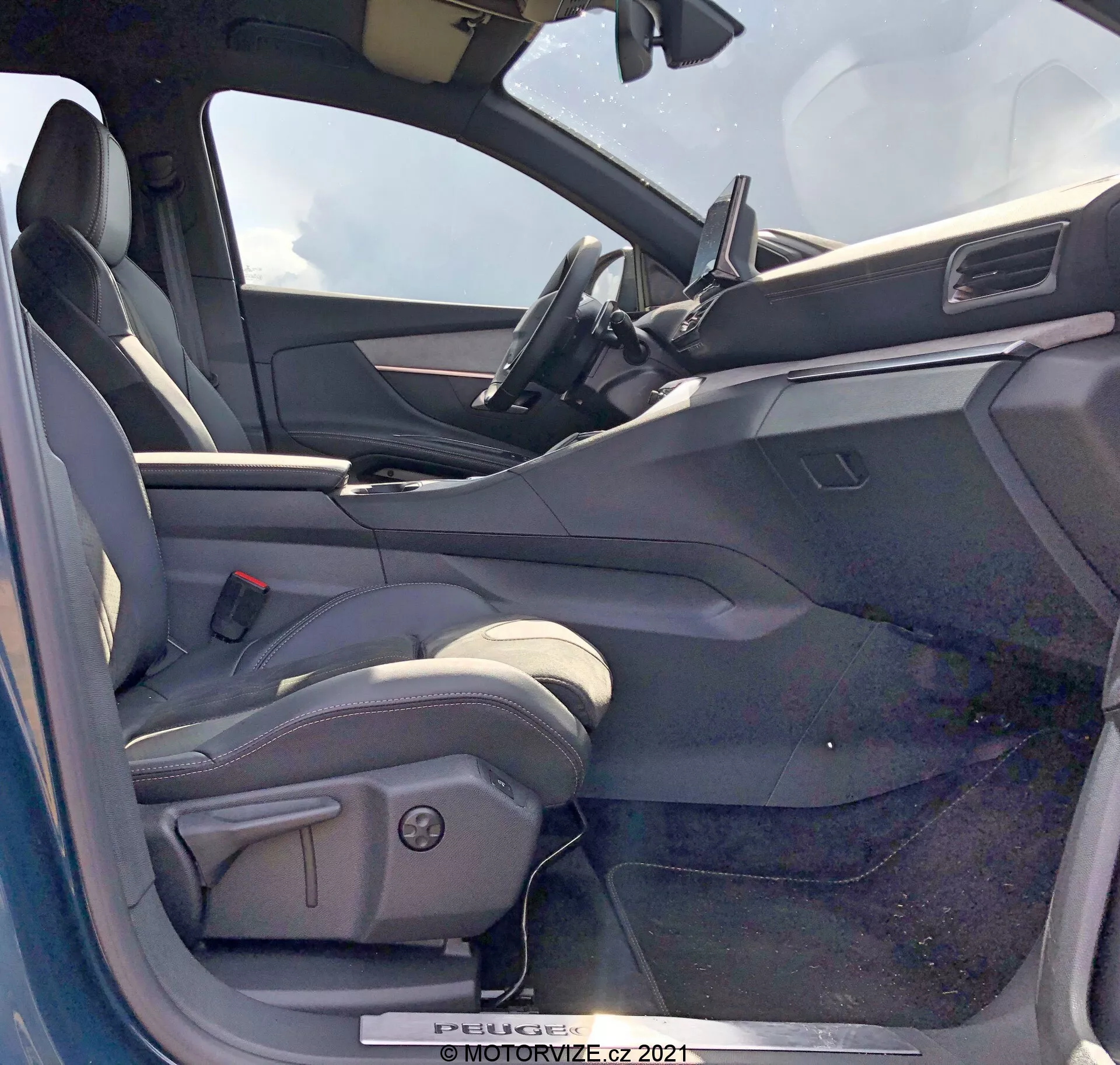 Vista del interior de un Peugeot 5008 de 2021 desde el lado del conductor con la puerta abierta. El asiento del conductor es de tipo bucket, tapizado probablemente en cuero o material similar al cuero, con refuerzos laterales y controles de ajuste eléctrico. La puerta del lado del conductor abierta muestra un panel interior con controles de ventanillas eléctricas, posiblemente controles de espejos y mecanismos de cierre. El salpicadero incluye una pantalla táctil de infoentretenimiento situada en el centro, con un diseño minimalista. La parte superior del volante es visible, sugiriendo un volante forrado en cuero con controles integrados. El interior tiene un esquema de colores de dos tonos con superficies superiores más oscuras y superficies inferiores más claras.