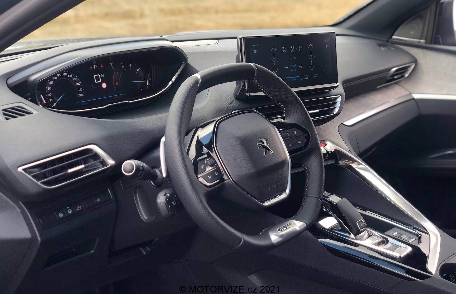 Vista interior del Peugeot 5008 2021 desde la perspectiva del conductor, mostrando el volante con el logo de Peugeot, múltiples botones de control, un cuadro de instrumentos completamente digital, pantalla de infoentretenimiento central con mapa de navegación, tablero elegante con botones físicos mínimos, consola central con botones para control de clima y configuraciones del vehículo, palanca de cambios electrónica, rejillas de ventilación, botón de arranque/parada del motor, materiales de gama alta y vista parcial del reposacabezas del asiento del conductor.