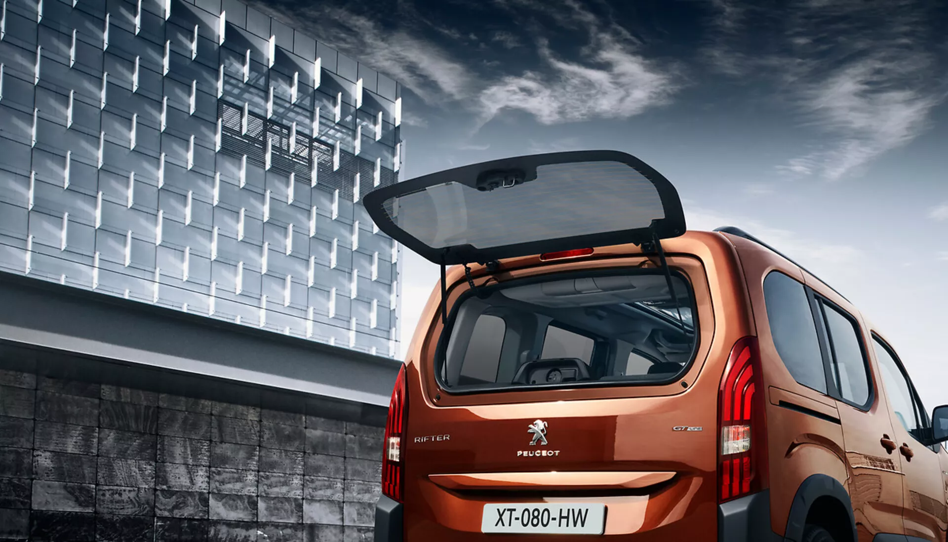 Vista trasera angular del Peugeot Rifter 2018 mostrando su diseño hatchback, acabado metálico, portón trasero abierto con espacio de carga visible, luces traseras LED y telón de fondo urbano.
