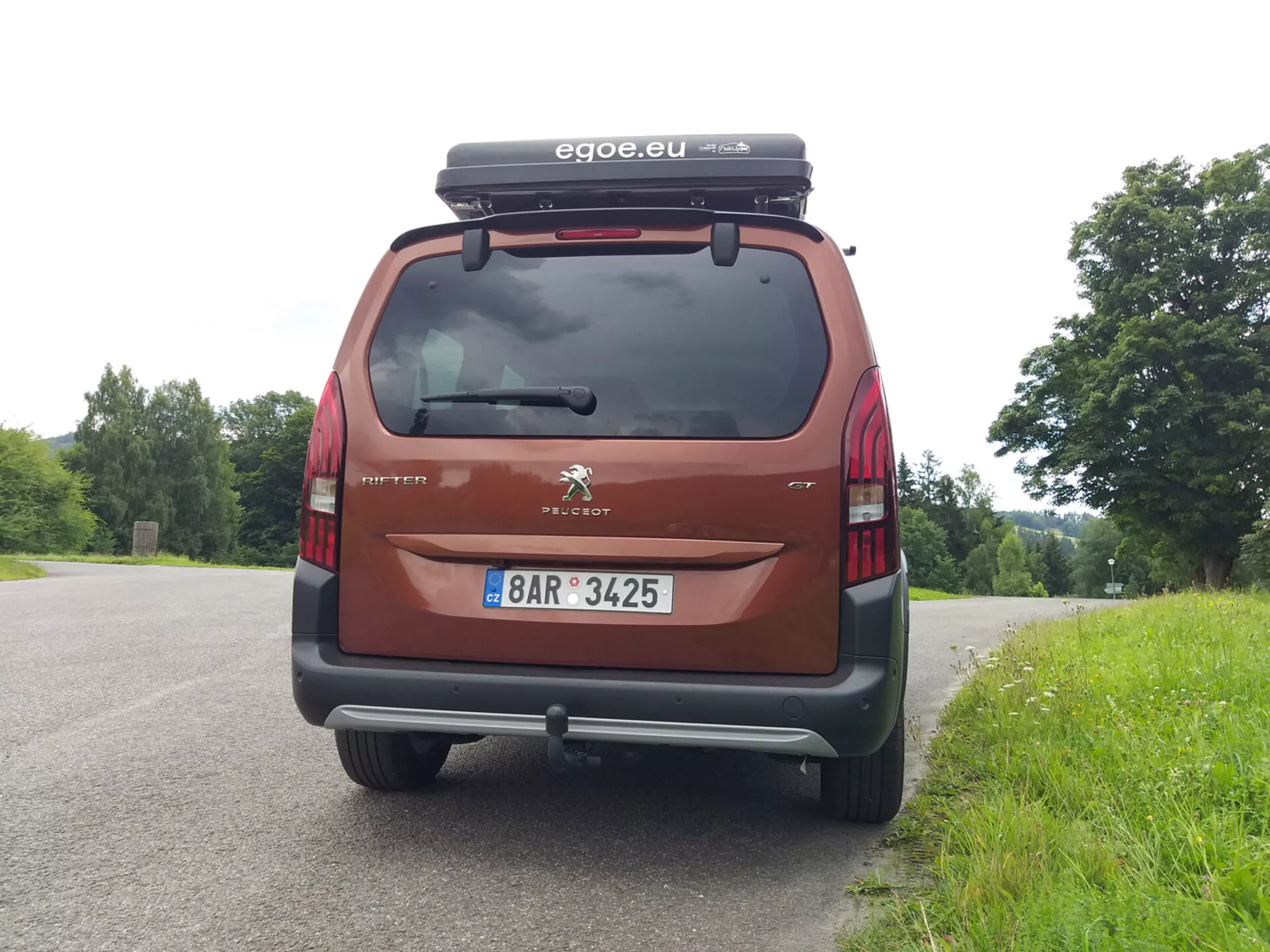 Vista trasera de un Peugeot Rifter 2018, mostrando el parabrisas trasero, las luces traseras, el parachoques y la matrícula, con el logo de Peugeot y el nombre del modelo Partner visibles. El coche, aparcado contra un telón de fondo natural, cuenta con un cofre de techo y lleva un accesorio de la marca 'egoe.eu', en color rojo/granate.