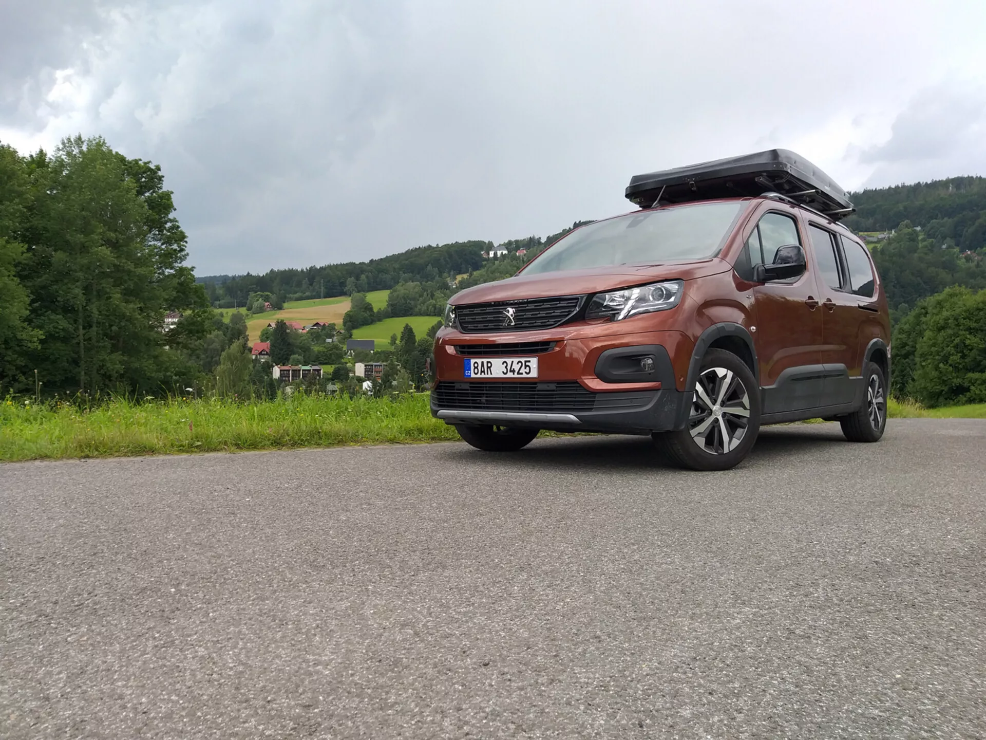 SUV Peugeot Rifter 2018 con pintura metálica y caja de techo capturada desde un ángulo bajo enfatizando la parrilla, el parachoques y las ruedas de aleación, mostrando capacidades todoterreno y diseño moderno incluyendo faros LED y sistemas de asistencia al conductor ADAS, estacionado en una superficie pavimentada contra un paisaje rural
