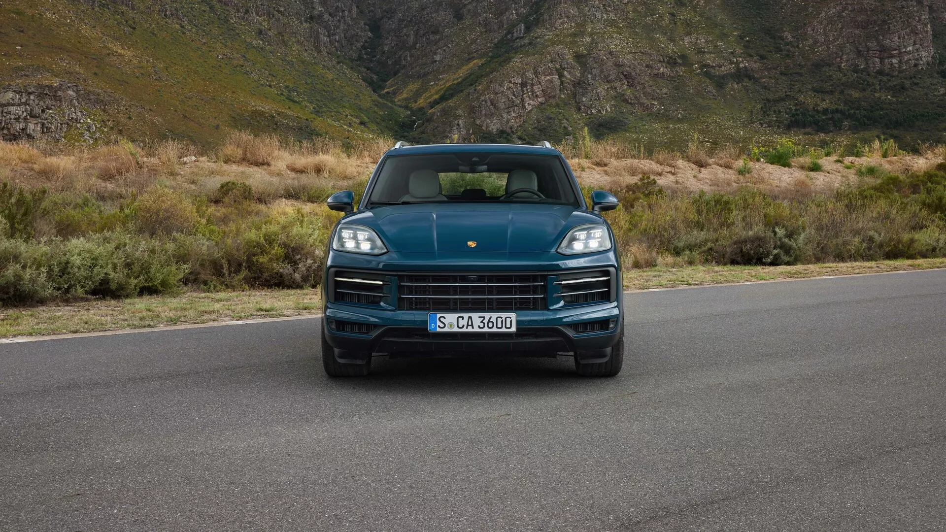 Porsche Cayenne 2023 (9YA Facelift) en una vista frontal a nivel de los ojos mostrando los faros LED, la parrilla prominentemente expuesta con el logotipo de la marca, diseño deportivo del capó y sensores de aparcamiento integrados, contra un telón de fondo montañoso indicando lujo y aventura