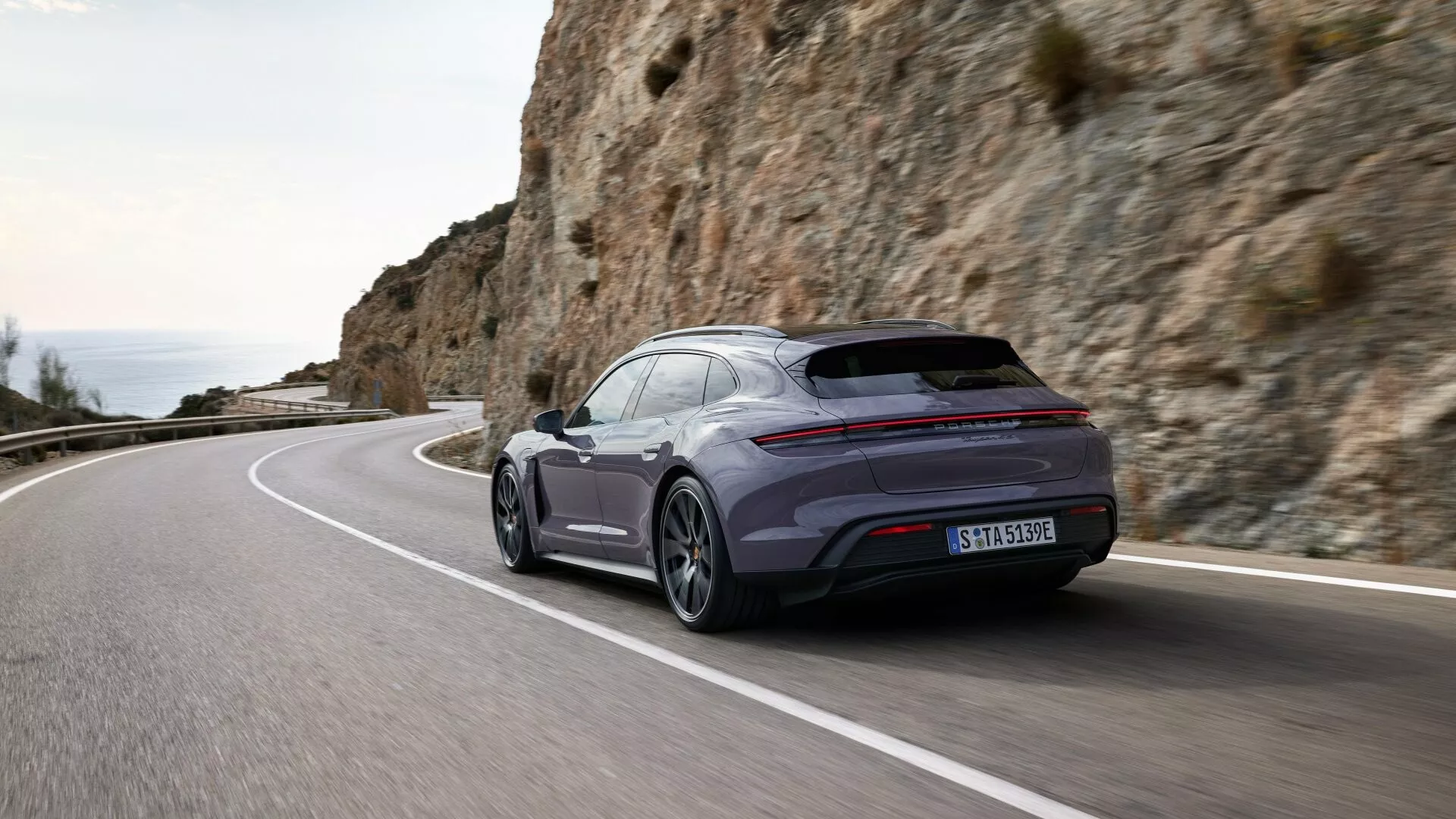 Vista trasera dinámica de tres cuartos del Porsche Taycan 2024 Y1A facelift en una carretera costera, destacando la postura deportiva, silueta aerodinámica y características tecnológicas avanzadas.