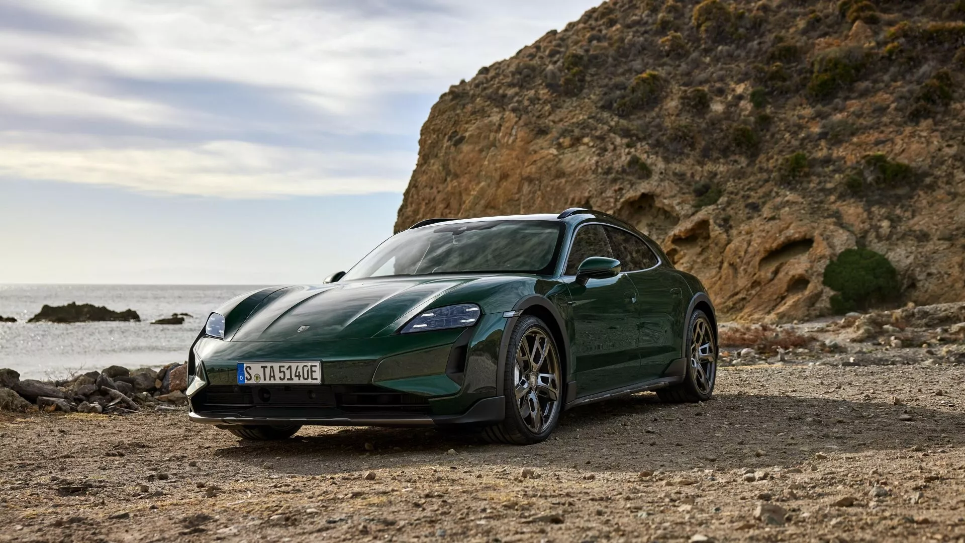 Modelo Porsche Taycan Y1A facelift 2024 fotografiado a la luz del día en un entorno costero sin pavimentar, mostrando su pintura verde oscuro metálica, diseño aerodinámico elegante y características avanzadas de vehículo eléctrico contra un telón de fondo de rocas y mar.