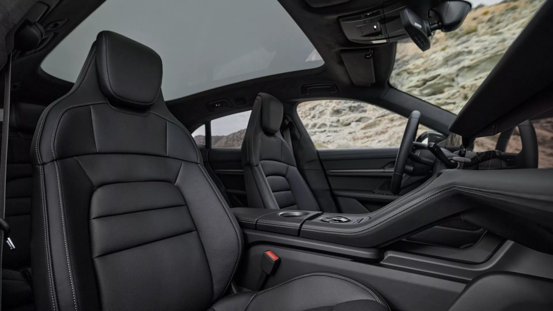 Modelo 2024 de Porsche Taycan Y1A facelift mostrando vista interior de lujo del coche desde los asientos traseros, centrándose en el asiento delantero de pasajero de cuero negro premium, consola central elegante, e indicios de diseño avanzado del tablero e interior de la puerta con techo convertible retraído, enfatizando la artesanía, la comodidad y las características tecnológicas.