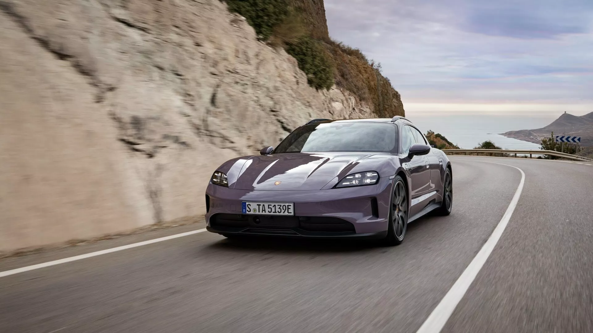 Porsche Taycan Y1A reestilización 2024 conduciendo por una carretera costera europea con una vista dinámica de tres cuartos delanteros que muestra una postura agresiva, diseño aerodinámico, faros LED, ruedas de aleación y neumáticos de rendimiento bajo la brillante luz del día