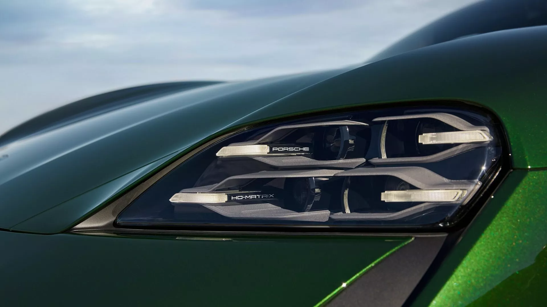 Vista detallada del faro del Porsche Taycan 2024 (restyling Y1A), mostrando tecnología de iluminación avanzada con elementos LED, marca PORSCHE y un vistazo al capó y guardabarros verde metálico que reflejan una aerodinámica sofisticada.