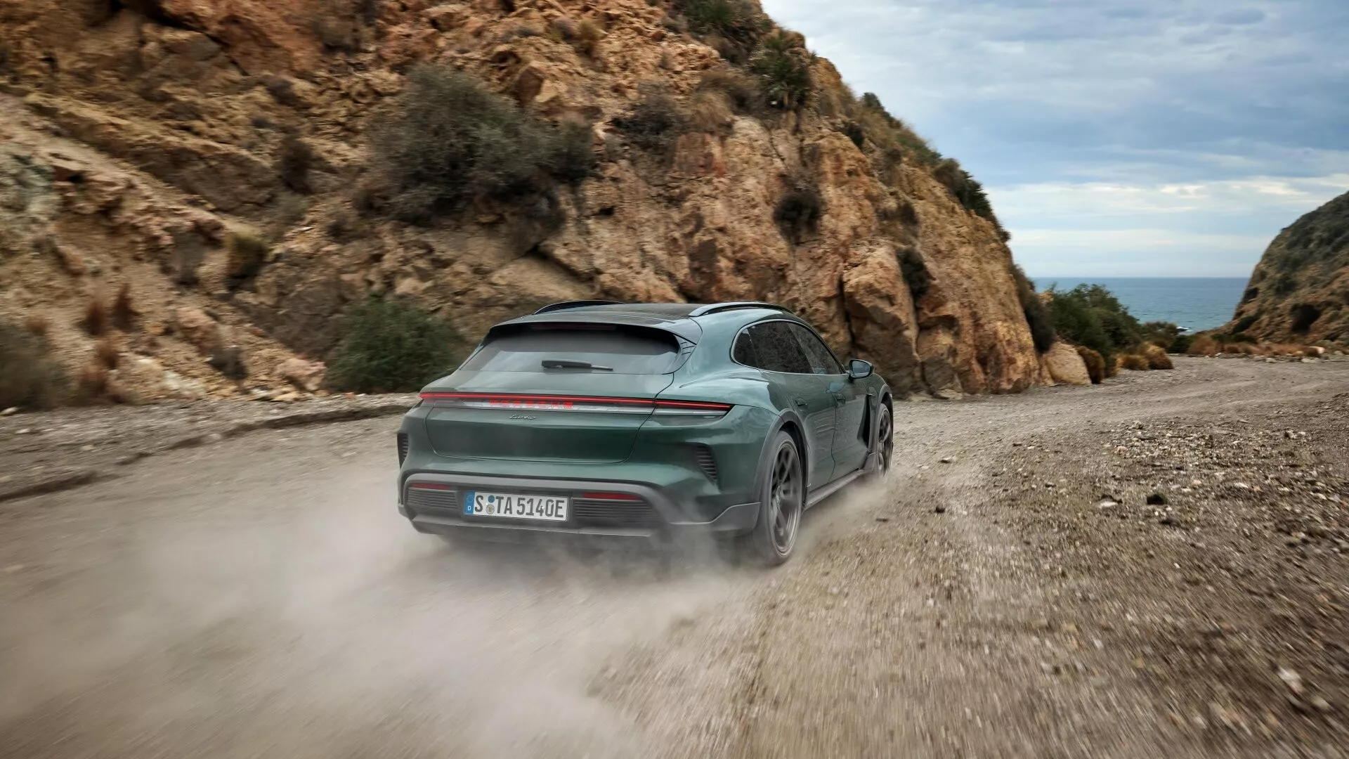 Porsche Taycan 2024 en verde, mostrando su diseño dinámico desde una vista trasera lateral en un camino de grava, con una distintiva barra de luces trasera y carrocería de dos tonos, insinuando capacidades de tracción total y tren motriz eléctrico, en medio de un paisaje natural rocoso.