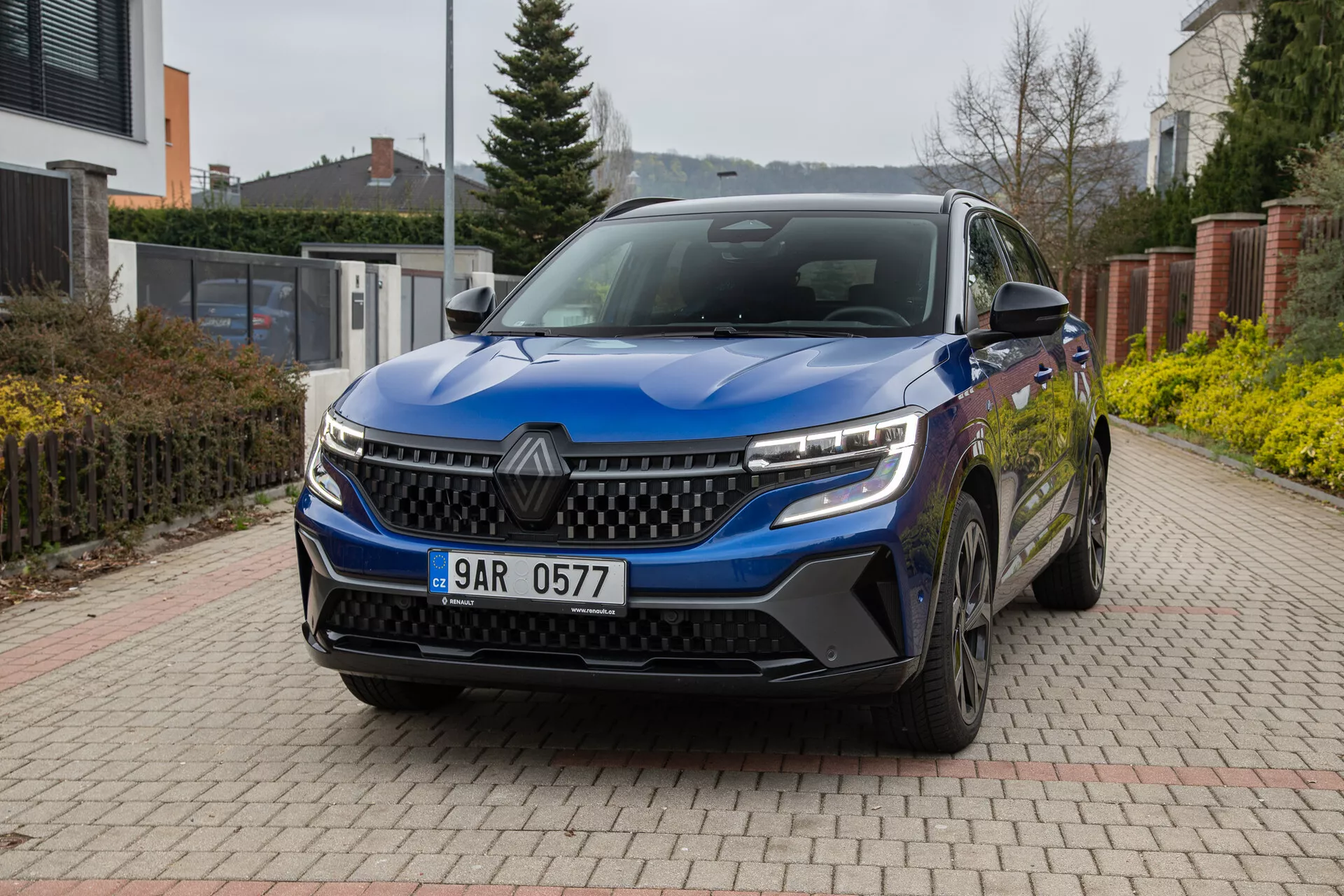 Renault Austral 2022 en azul, mostrando su ángulo frontal de tres cuartos en un camino pavimentado con ladrillos con entorno residencial al fondo, destacando faros LED, parrilla distintiva, sensores frontales, ruedas de aleación y espejos laterales con señales de giro integradas bajo luz natural y nublada.