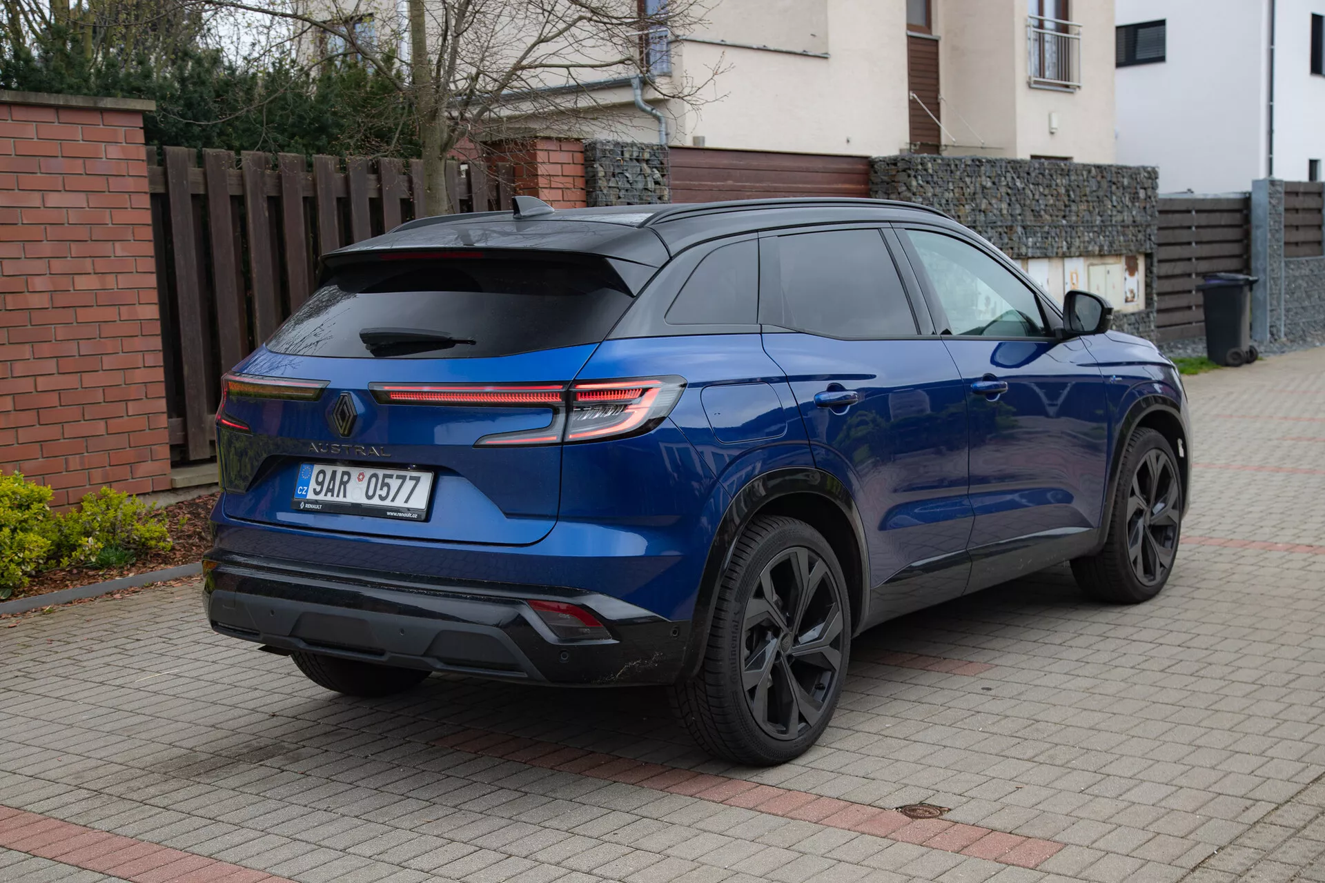SUV moderno Renault Austral 2022 en azul metálico brillante estacionado en una entrada pavimentada al lado de un edificio residencial, mostrando su spoiler trasero deportivo, luces traseras LED, ruedas de aleación de color oscuro, ventanas traseras tintadas y sensores de aparcamiento desde un ángulo trasero de tres cuartos