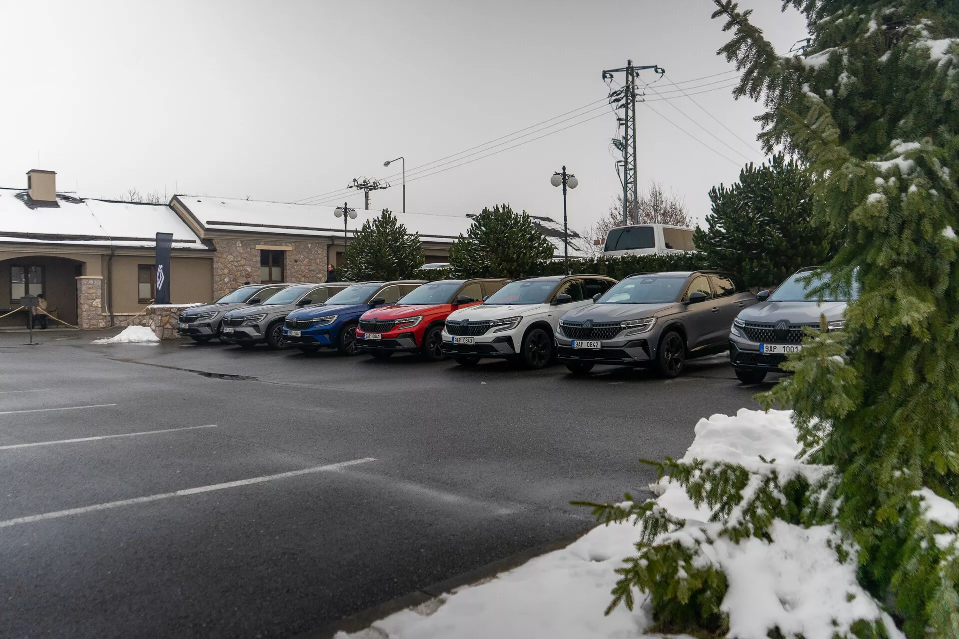 Fila de autos estacionados de sedanes y SUVs bajo una ligera nevada, indicando clima frío, con perfiles laterales visibles incluyendo puertas, espejos laterales y ruedas, tomada desde un ángulo elevado de la acera