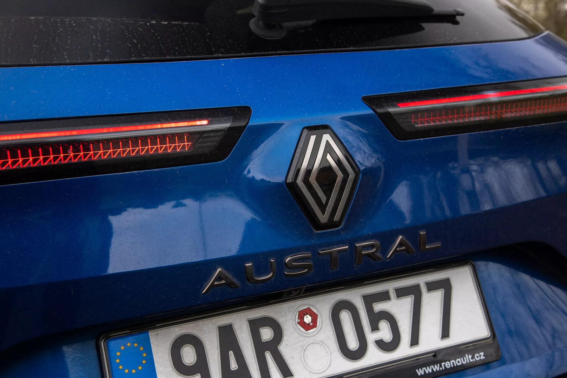 Imagen detallada de la sección trasera del Renault Austral 2022 en azul brillante, con logo diamante centrado y nombre del modelo 'Austral' en letras mayúsculas sans-serif, placa de matrícula con bandera de la Unión Europea, distintivos faros traseros LED horizontales y posible sistema adicional de iluminación trasera, exterior del coche indica uso reciente.