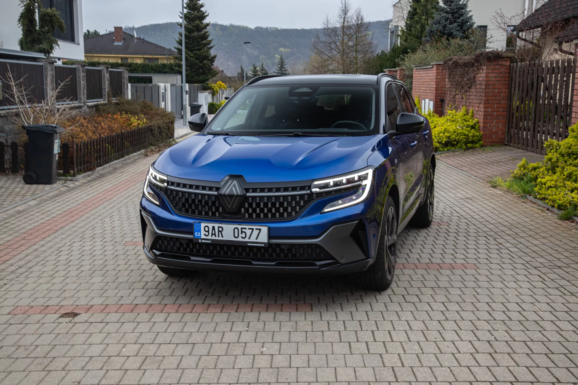 Vista frontal en ángulo de un SUV Renault Austral 2022 estacionado en un camino residencial, mostrando la parrilla, faros LED, luces diurnas, parachoques con tomas de aire, sensores de estacionamiento, emblema, ruedas de aleación, espejos laterales con señales de giro y placa de matrícula.
