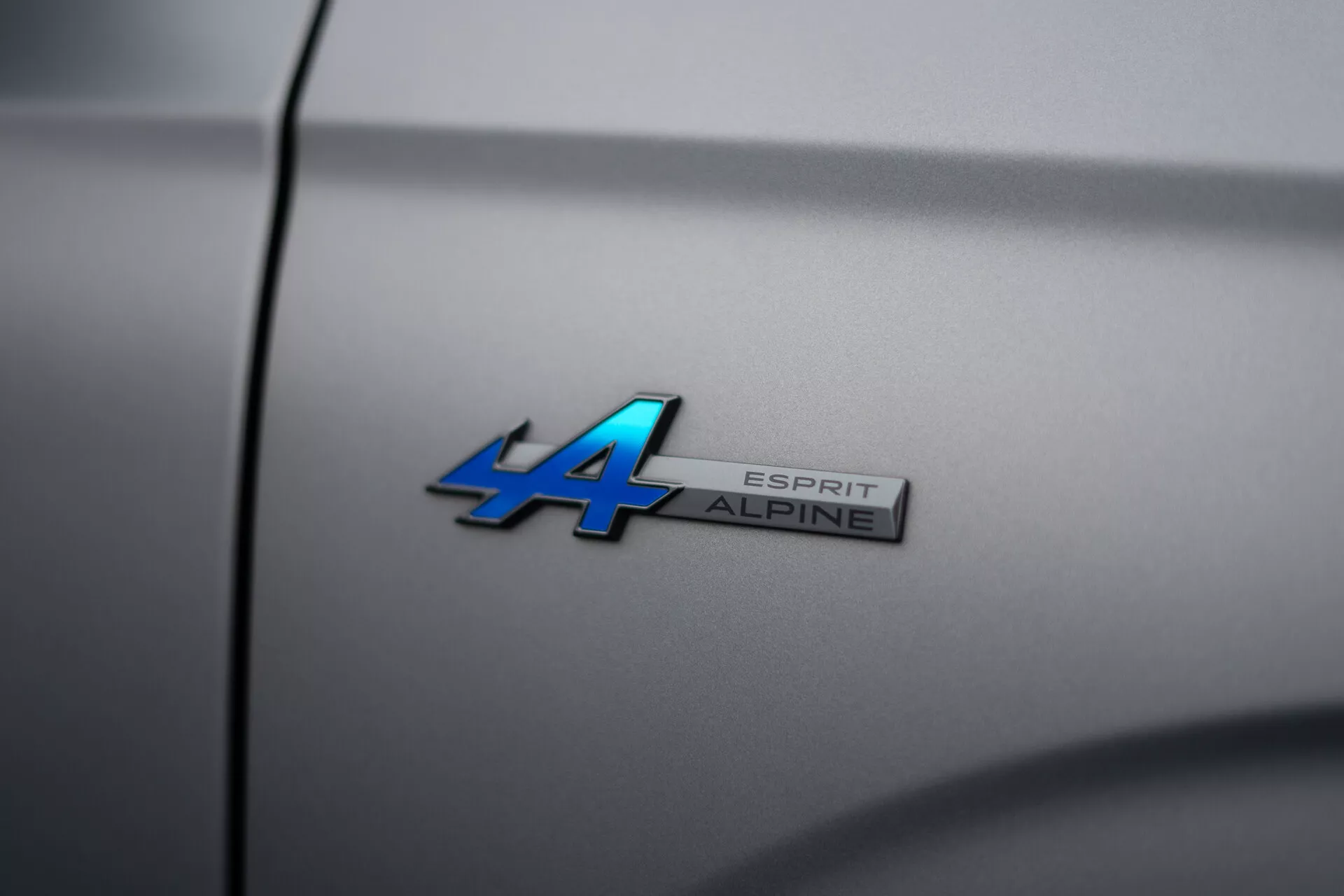 Imagen de primer plano del emblema de Alpine en el Renault Austral 2022 con designación de modelo 'ALPINE ESPRIT' en superficie metálica azul y cromo, angulada para profundidad de campo reducida