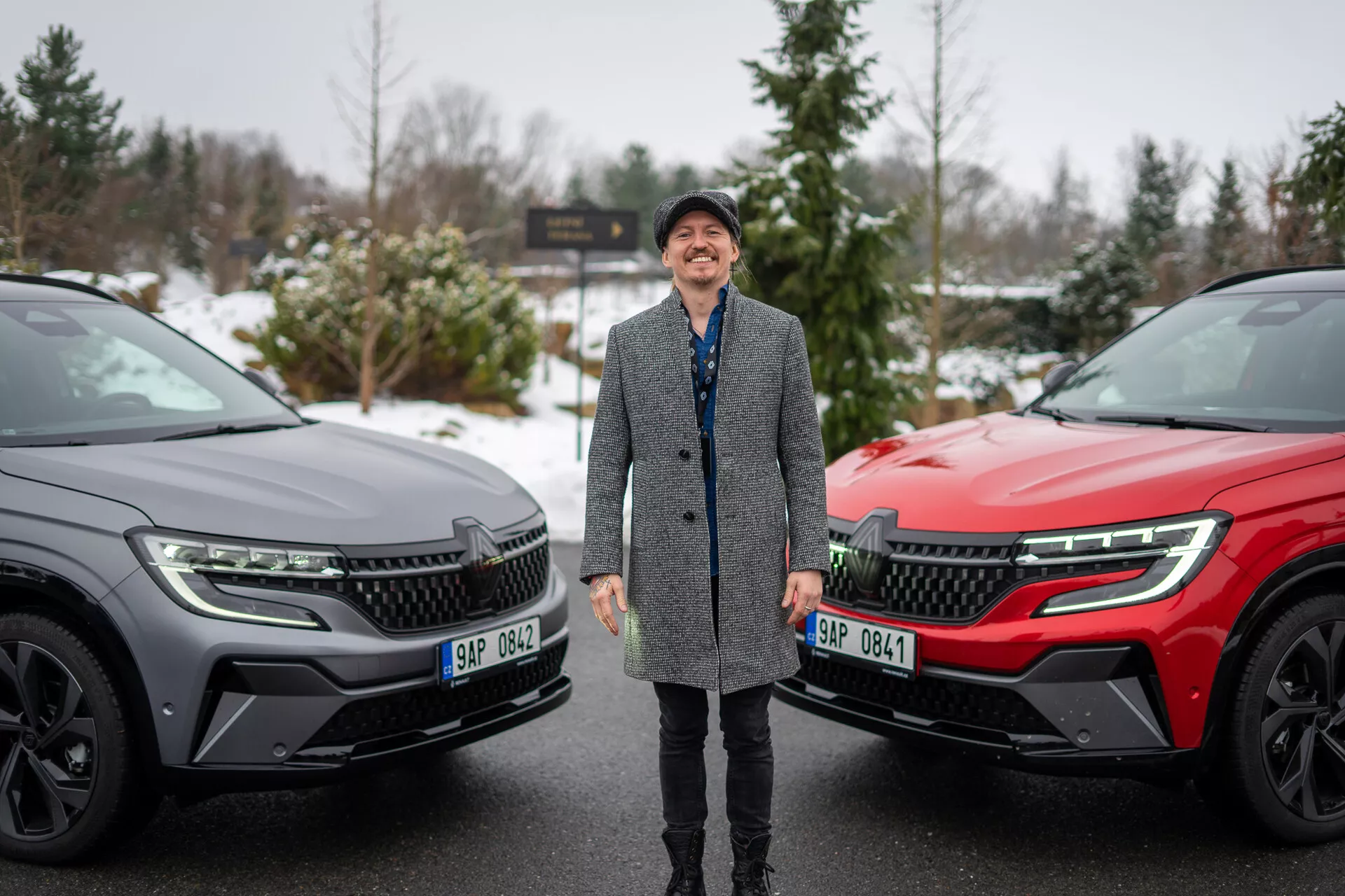 Persona sonriente flanqueada por dos SUV Renault Austral 2022 en un entorno invernal, destacando el diseño moderno de los vehículos y las características tecnológicas avanzadas con foco en la parrilla frontal, faros LED y perfil lateral elegante.