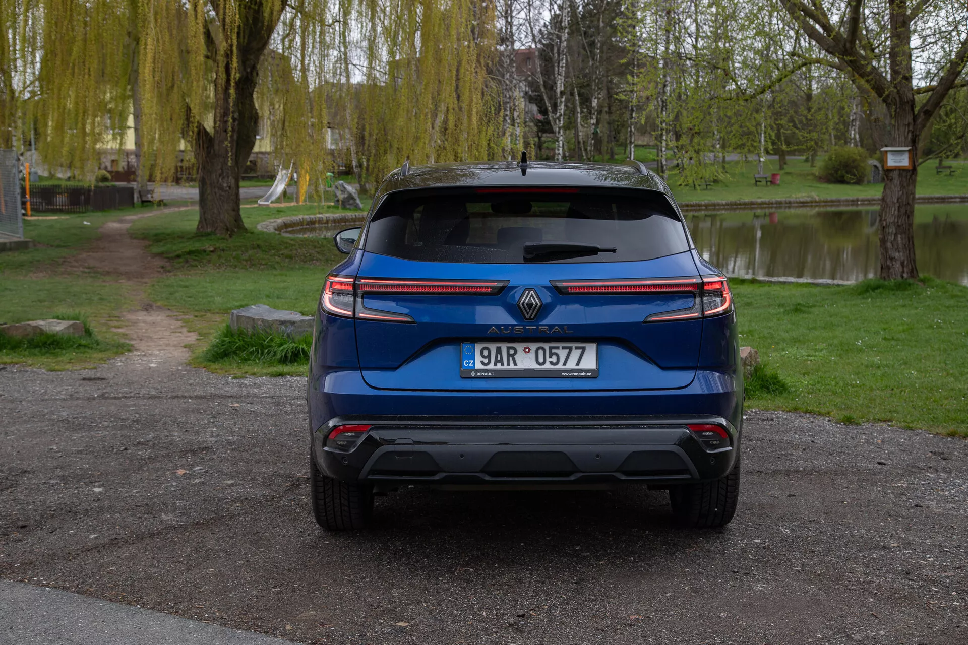 Vista trasera de un Renault Austral 2022 azul, aparcado al aire libre mostrando características que incluyen el logotipo de la marca, matrícula, luces traseras modernas, parabrisas trasero con limpiaparabrisas, parachoques trasero diseñado con posibles sensores de asistencia de aparcamiento, puntas de escape visibles, reflectores o sensores, y un alerón trasero para mejorar la aerodinámica, todo ello ambientado en un tranquilo entorno parecido a un parque con árboles, césped y un cuerpo de agua.