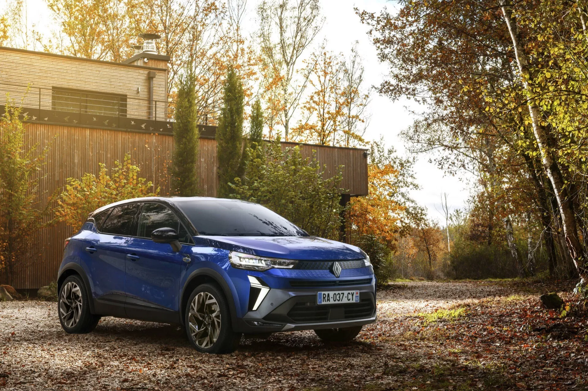 Renault Captur II facelift 2024 en azul, un SUV compacto aparcado al aire libre mostrando su vista del cuarto frontal derecho con un enfoque en sus líneas de diseño moderno, parrilla frontal distintiva, faros LED y ruedas de aleación, contra un telón de fondo otoñal bajo luz natural de mañana o tarde lo que realza su atractivo estético y avances tecnológicos