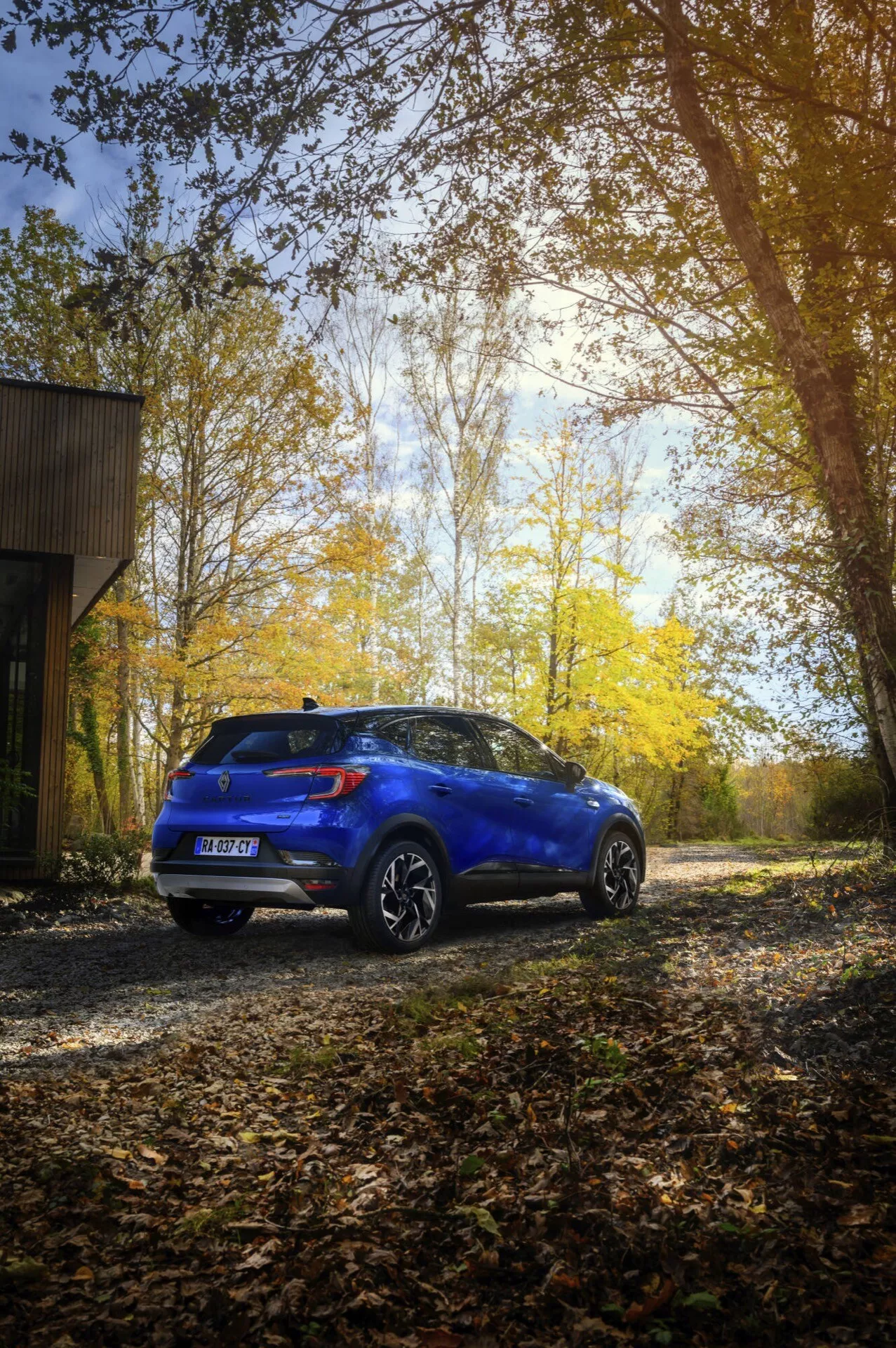 Modelo Renault Captur II facelift de 2024 en azul, mostrando su perfil trasero y lateral al aire libre sobre una superficie de grava con un fondo boscoso otoñal, destacando sus líneas de diseño, acabado brillante, ruedas de aleación y alerón trasero.